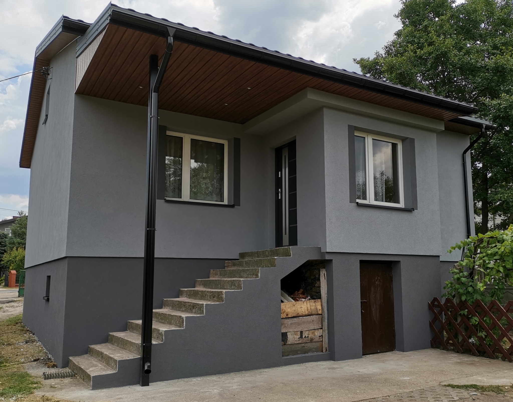 Dwukondygnacyjny dom z szarą elewacją, brązowym podbitką i schodami wejściowymi, z widocznym wejściem do piwnicy i drewnianym ogrodzeniem w tle.