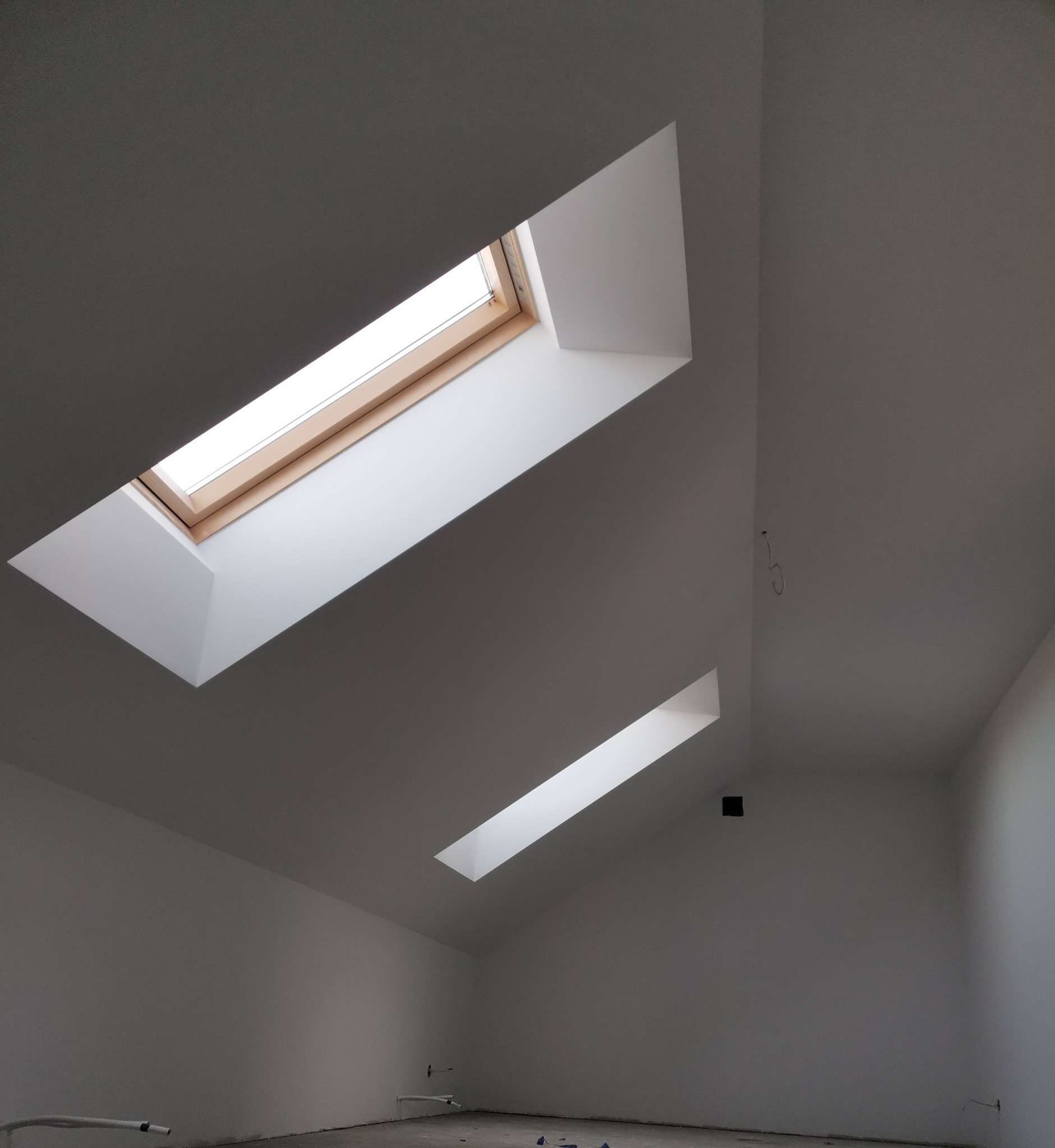 Poddasze z dwoma oknami dachowymi o drewnianych ramach i białych wnękach, świeżo pomalowane na biało, widoczne przewody i rury przy podłodze.