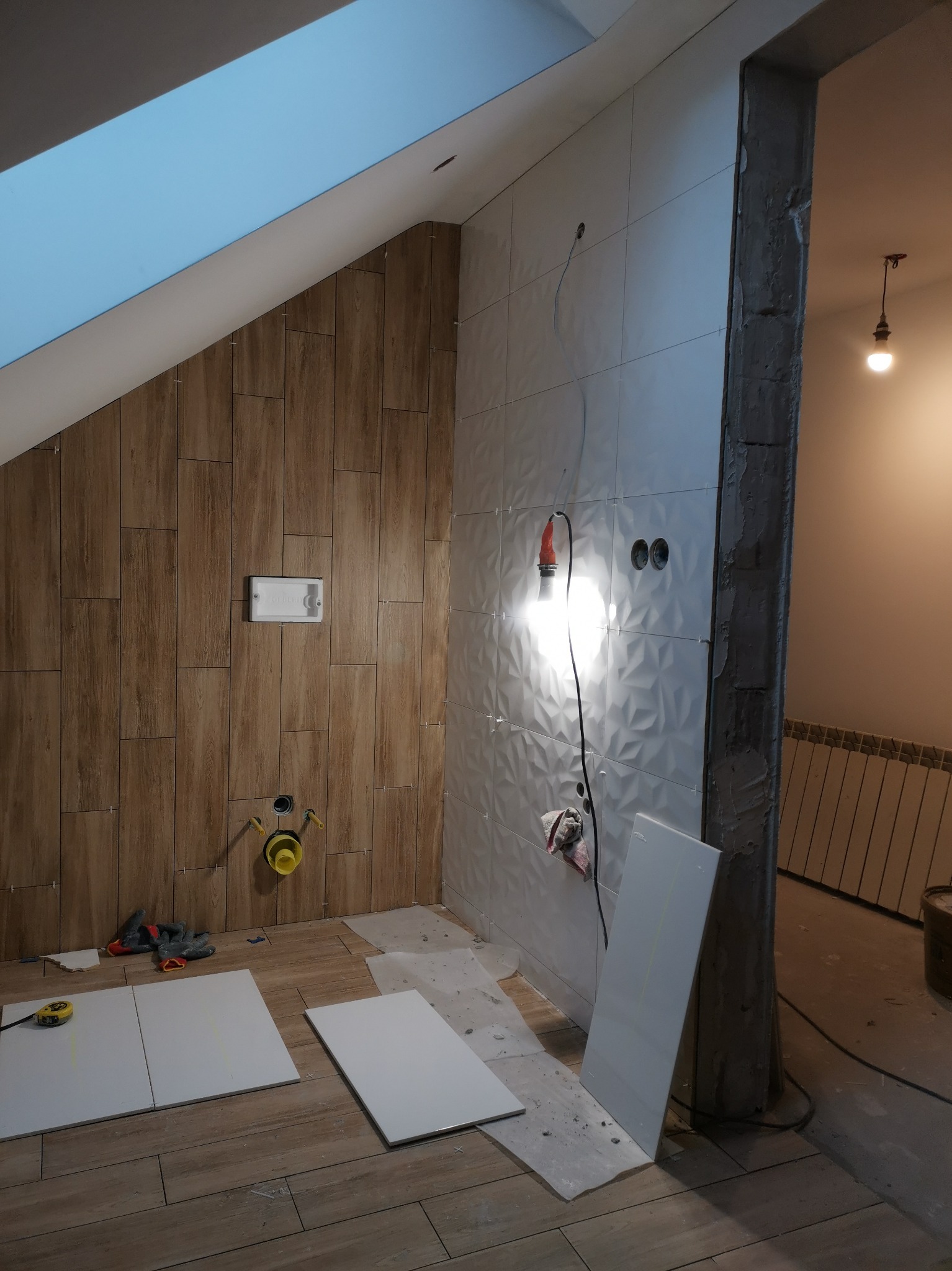 Wnętrze łazienki w trakcie remontu: częściowo ułożone drewnopodobne i białe płytki ścienne z wzorem 3D, widoczne narzędzia, przewody elektryczne i fragment sufitu skośnego z oknem dachowym.