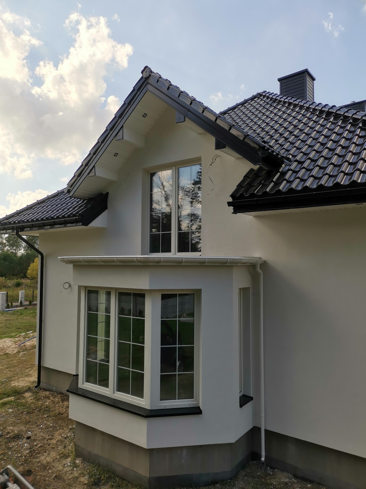 Widok z dołu na fragment otynkowanego, białego domu jednorodzinnego z czarną dachówką i wykuszem okiennym, z widocznym fragmentem niezagospodarowanego terenu.