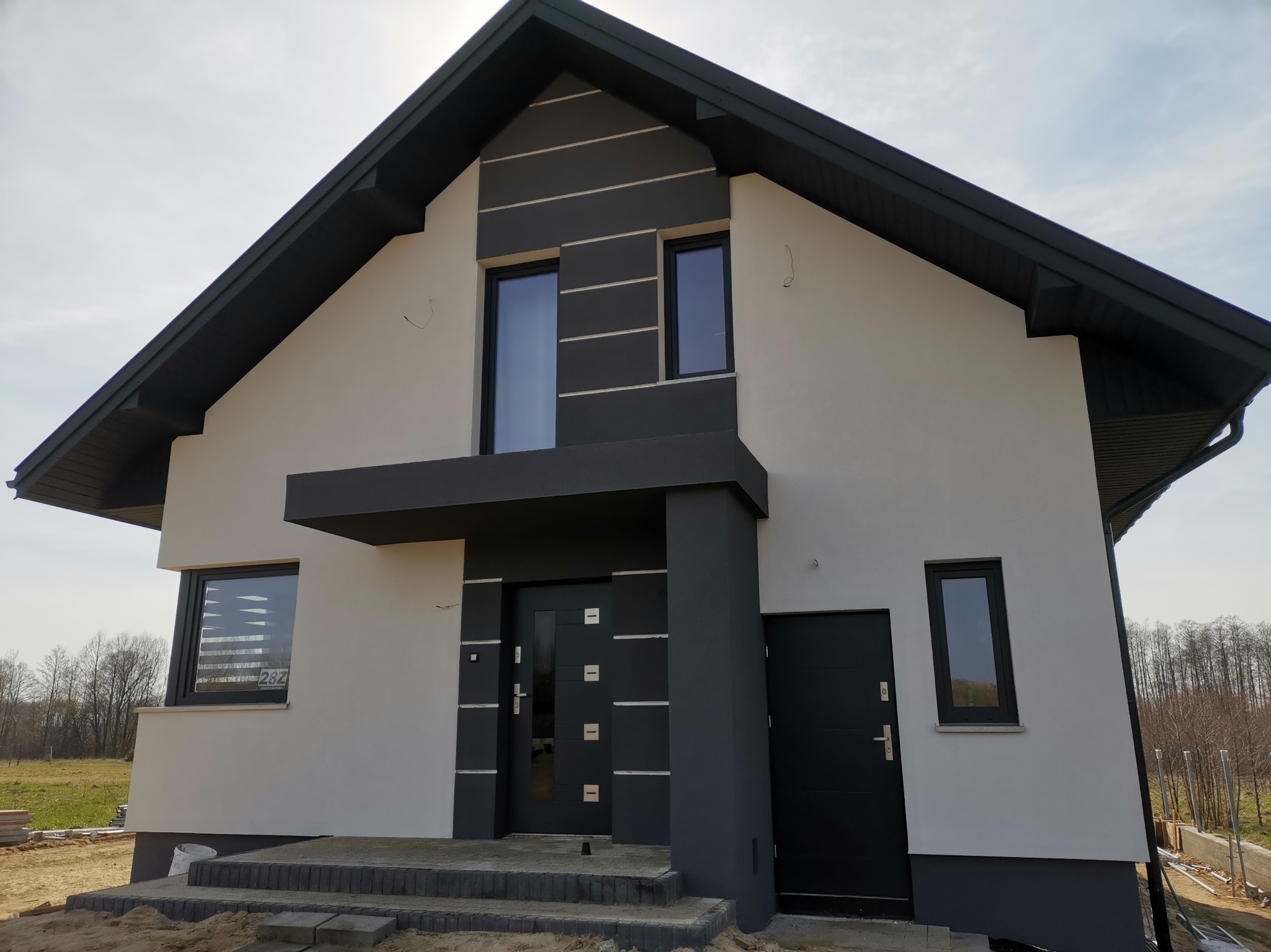 Nowoczesny dom jednorodzinny z elewacją w odcieniach szarości i bieli, widoczne schody wejściowe i teren w trakcie prac wykończeniowych.