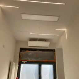 Nowoczesne oświetlenie LED liniowe na suficie w jasnym pomieszczeniu, widoczny klimatyzator i okno zabezpieczone folią malarską.