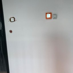 Elekrtoimpuls - Biała ściana z widocznymi elementami instalacji: otwór po gniazdku, otwór na kabel, czujnik oddymiania i panel sterowania PP-40.