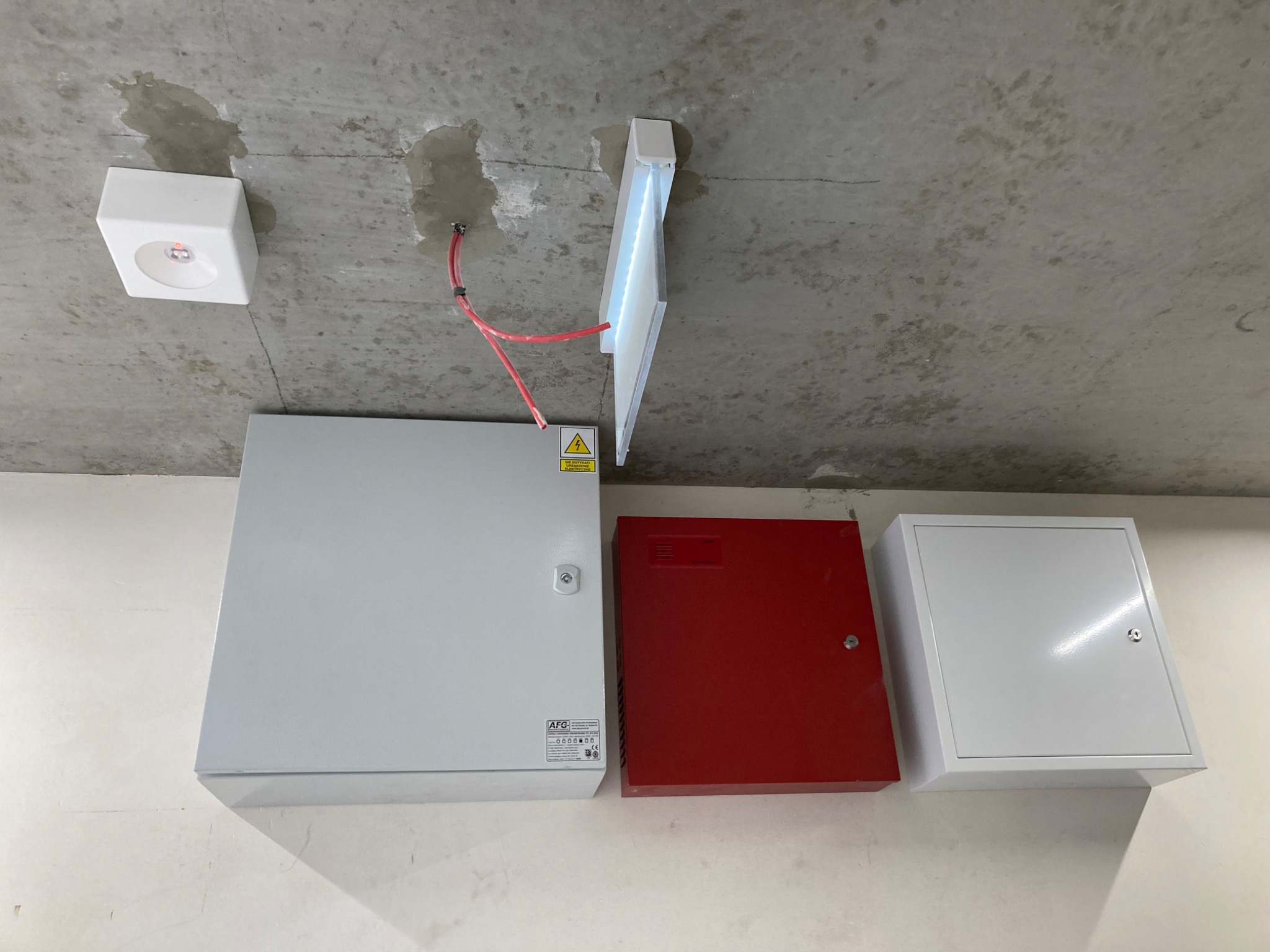 Na surowym betonowym suficie zamontowane trzy skrzynki: szara z naklejką ostrzegawczą, czerwona z zamkiem i biała z zamkiem, obok czujnik dymu i lampa LED z widocznymi kablami.