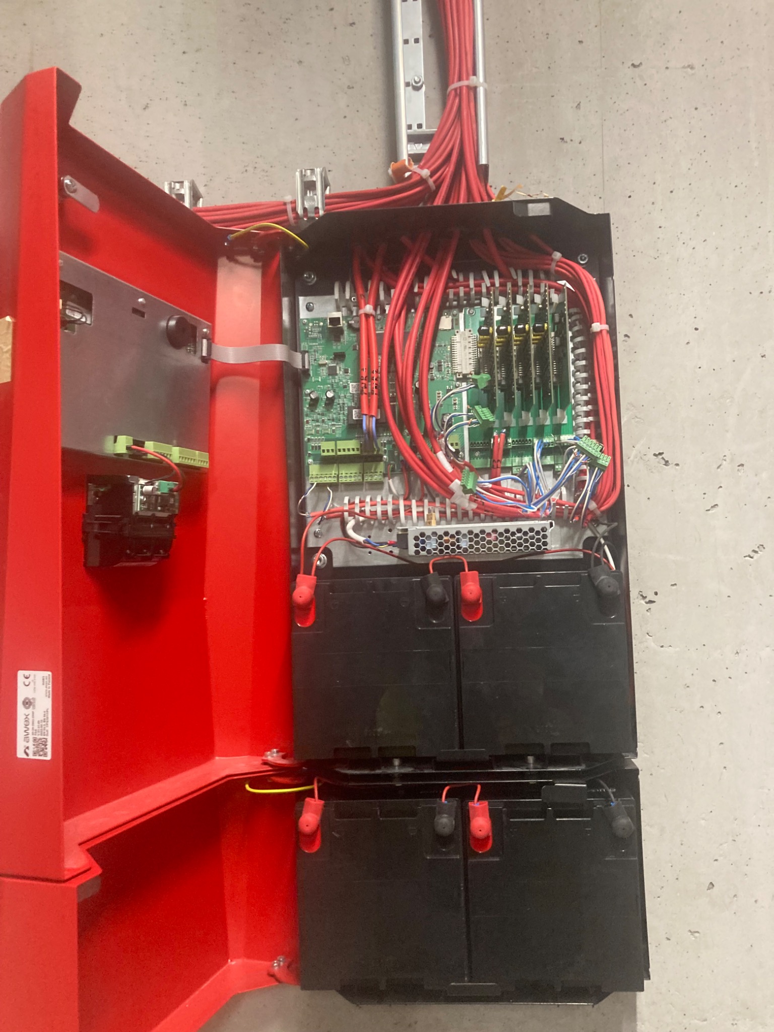 Otwarta szafka sterownicza systemu alarmowego z widocznymi płytkami drukowanymi, okablowaniem i akumulatorami, zamontowana na szarej ścianie.