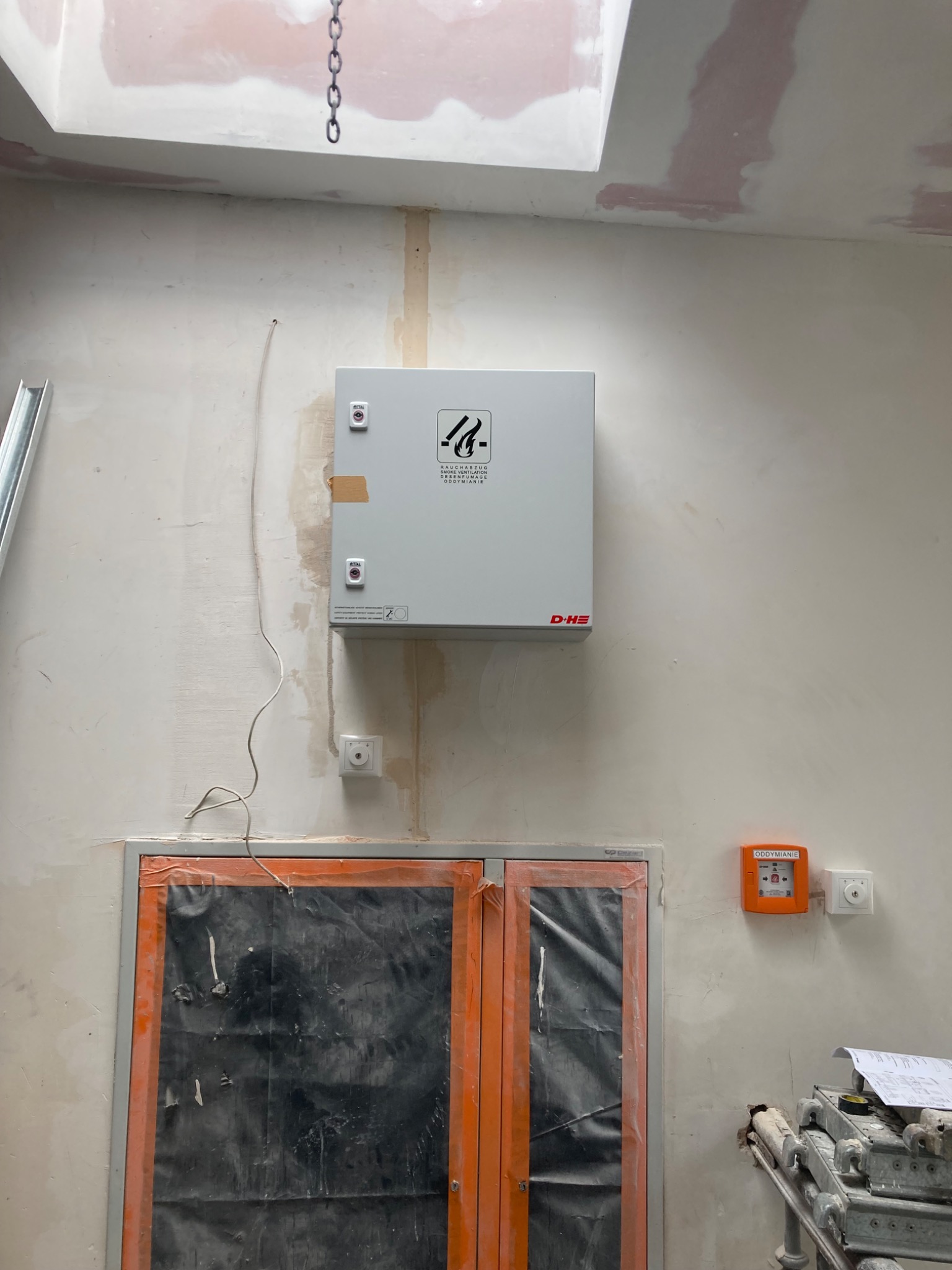 Biała skrzynka sterownicza systemu oddymiania zamontowana na ścianie w trakcie prac wykończeniowych, widoczny fragment drzwi zabezpieczonych folią, przycisk oddymiania i gniazdko elektryczne.