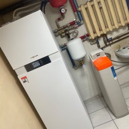 Hydro Tech - Nowoczesny, biały piec grzewczy Viessmann z wyświetlaczem, obok biały zmiękczacz wody Viteco z pomarańczową górną częścią, na tle starego, żeliwnego grzejnika i instalacji hydraulicznej.