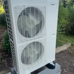 Biała pompa ciepła Viessmann Vitocal 100 umieszczona na betonowych blokach, otoczona zielenią w ogrodzie.