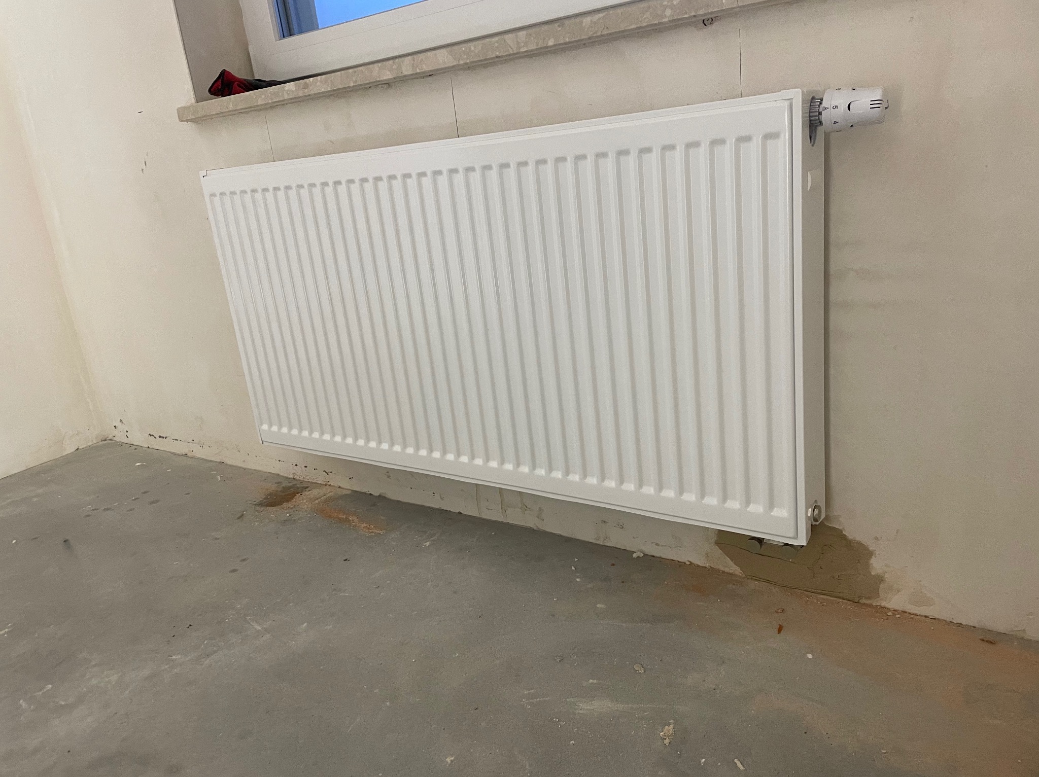 Biały, stalowy grzejnik panelowy z termostatem zamontowany na ścianie w surowym pomieszczeniu z betonową podłogą i śladami po zalaniu.