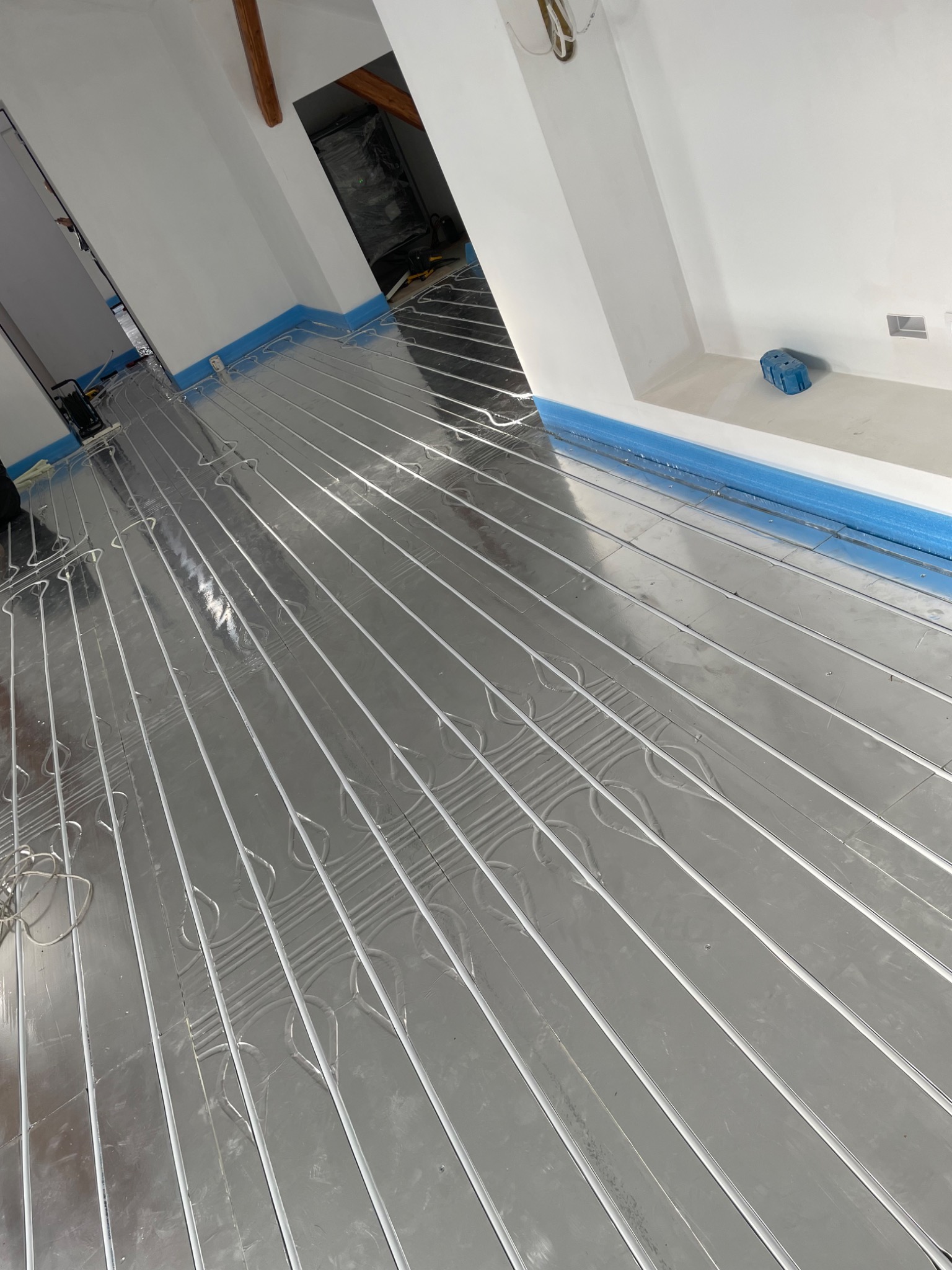 Białe rurki ogrzewania podłogowego ułożone na srebrnej folii izolacyjnej, widoczne niebieskie zabezpieczenia przy ścianach i fragment poddasza w tle.