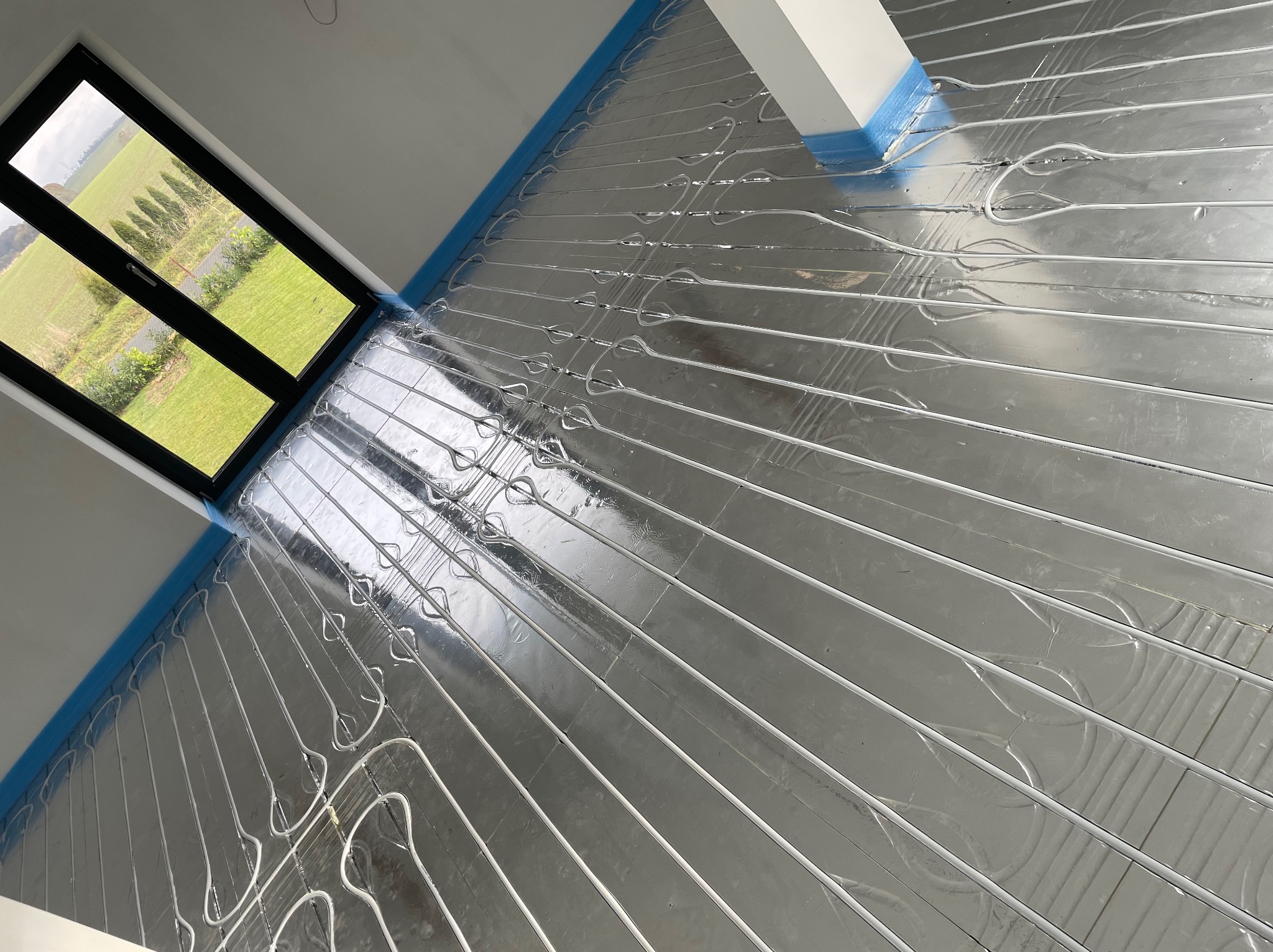 Instalacja ogrzewania podłogowego z widocznymi rurkami grzewczymi na srebrnej folii izolacyjnej, widok z góry, okno z widokiem na zielony krajobraz.