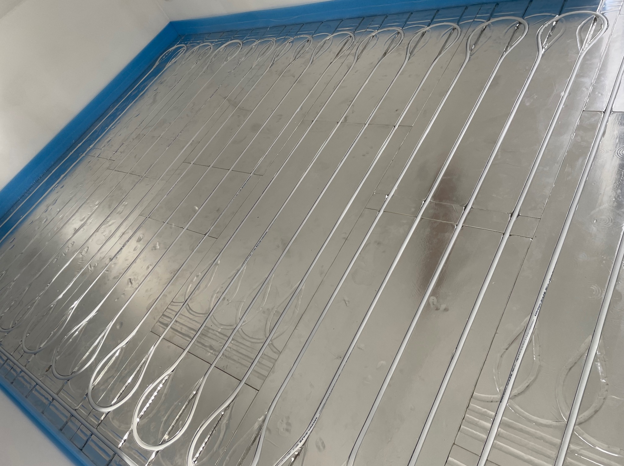 Rozłożona instalacja ogrzewania podłogowego na folii aluminiowej z niebieską taśmą brzegową, widok z góry.
