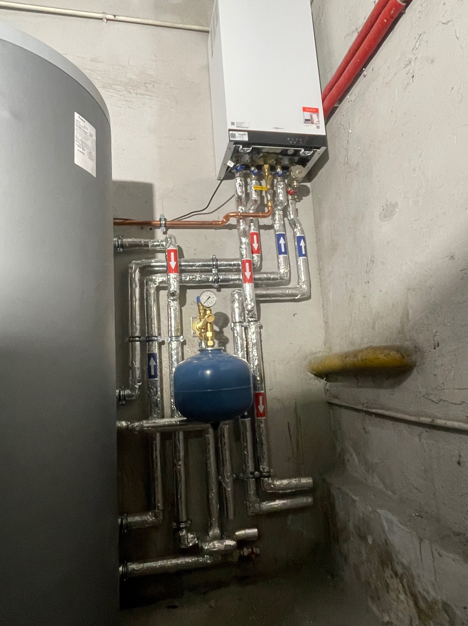 Zmontowany system grzewczy z kotłem, zbiornikiem akumulacyjnym i rurami z izolacją, strzałki na rurach wskazują kierunek przepływu, manometr na niebieskim naczyniu wzbiorczym.