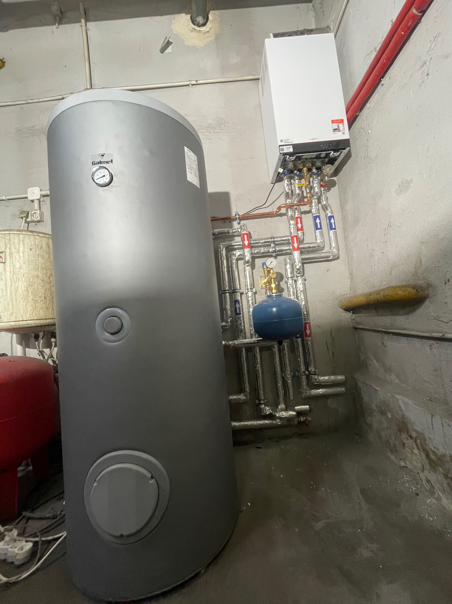 Instalacja grzewcza w piwnicy: szary zasobnik ciepłej wody Galmet, biały piec gazowy Viessmann, rury miedziane i aluminiowe z izolacją, manometr na niebieskim zbiorniku wyrównawczym, betonowe...