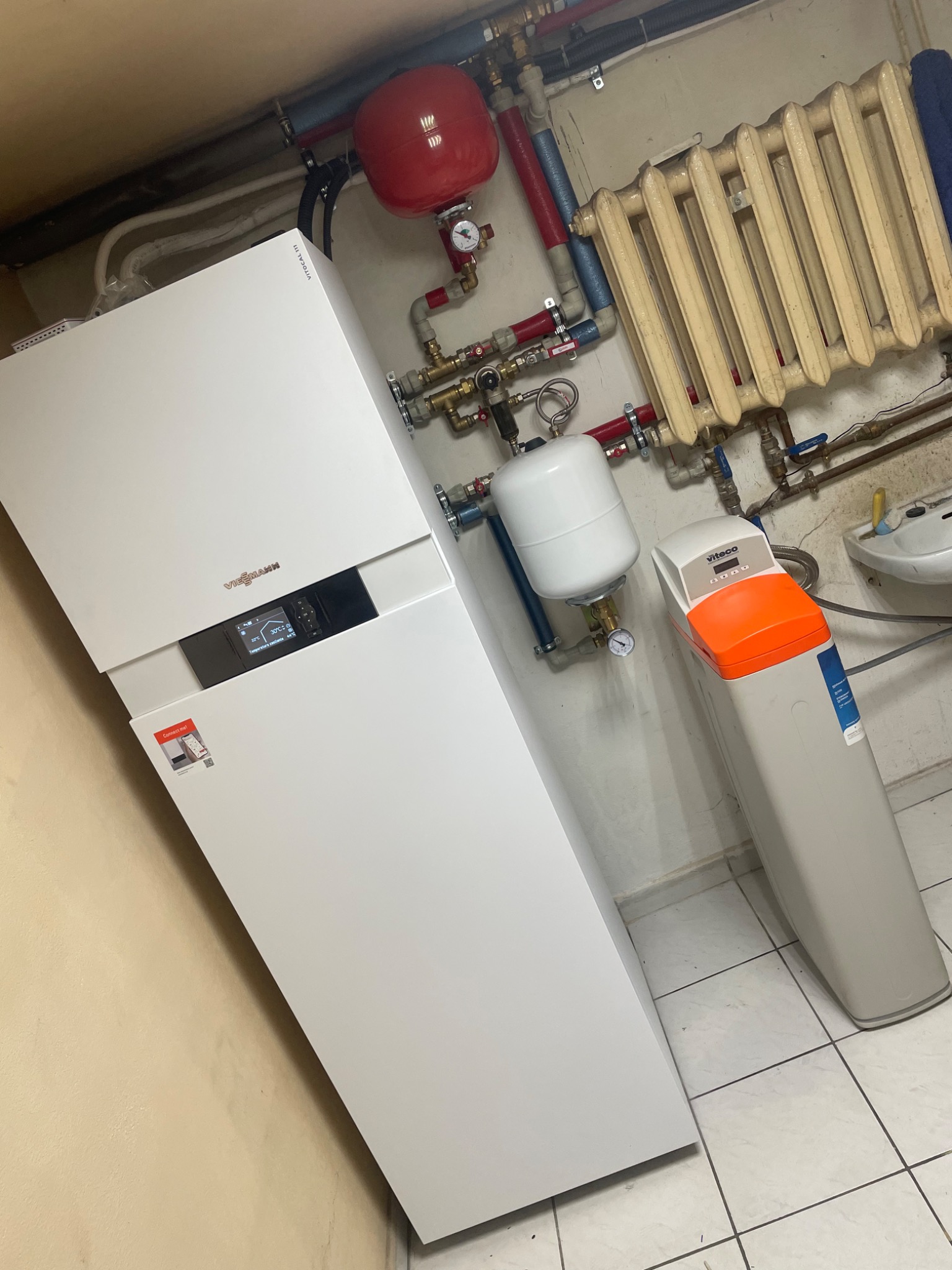 Nowoczesny, biały piec grzewczy Viessmann z wyświetlaczem, obok biały zmiękczacz wody Viteco z pomarańczową górną częścią, na tle starego, żeliwnego grzejnika i instalacji hydraulicznej.