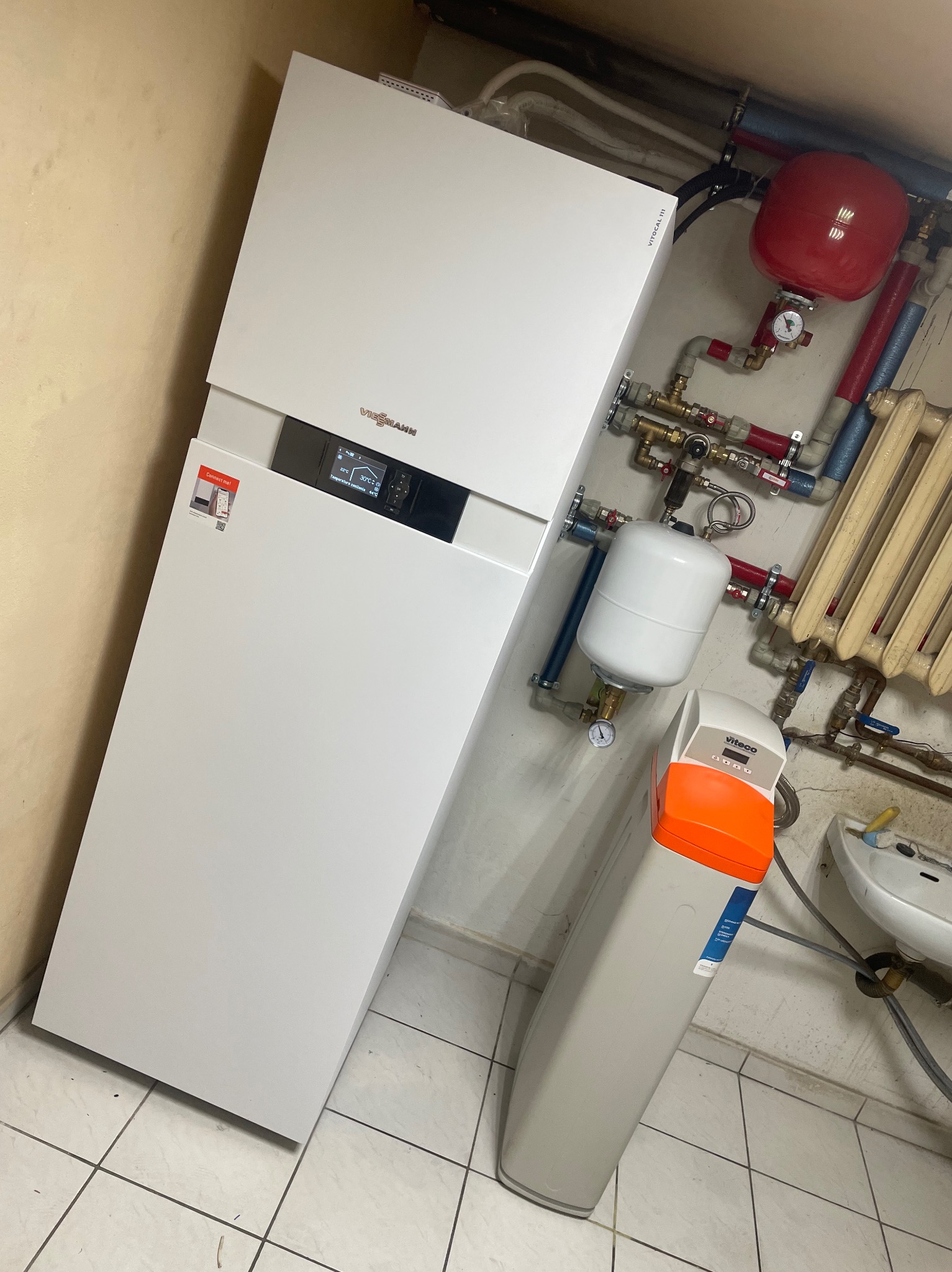 Kompaktowy, biały piec grzewczy Viessmann z widocznym wyświetlaczem, obok biały filtr wody Viteco z pomarańczową górną częścią, czerwony zbiornik wyrównawczy i fragment grzejnika żeberkowego w tle.