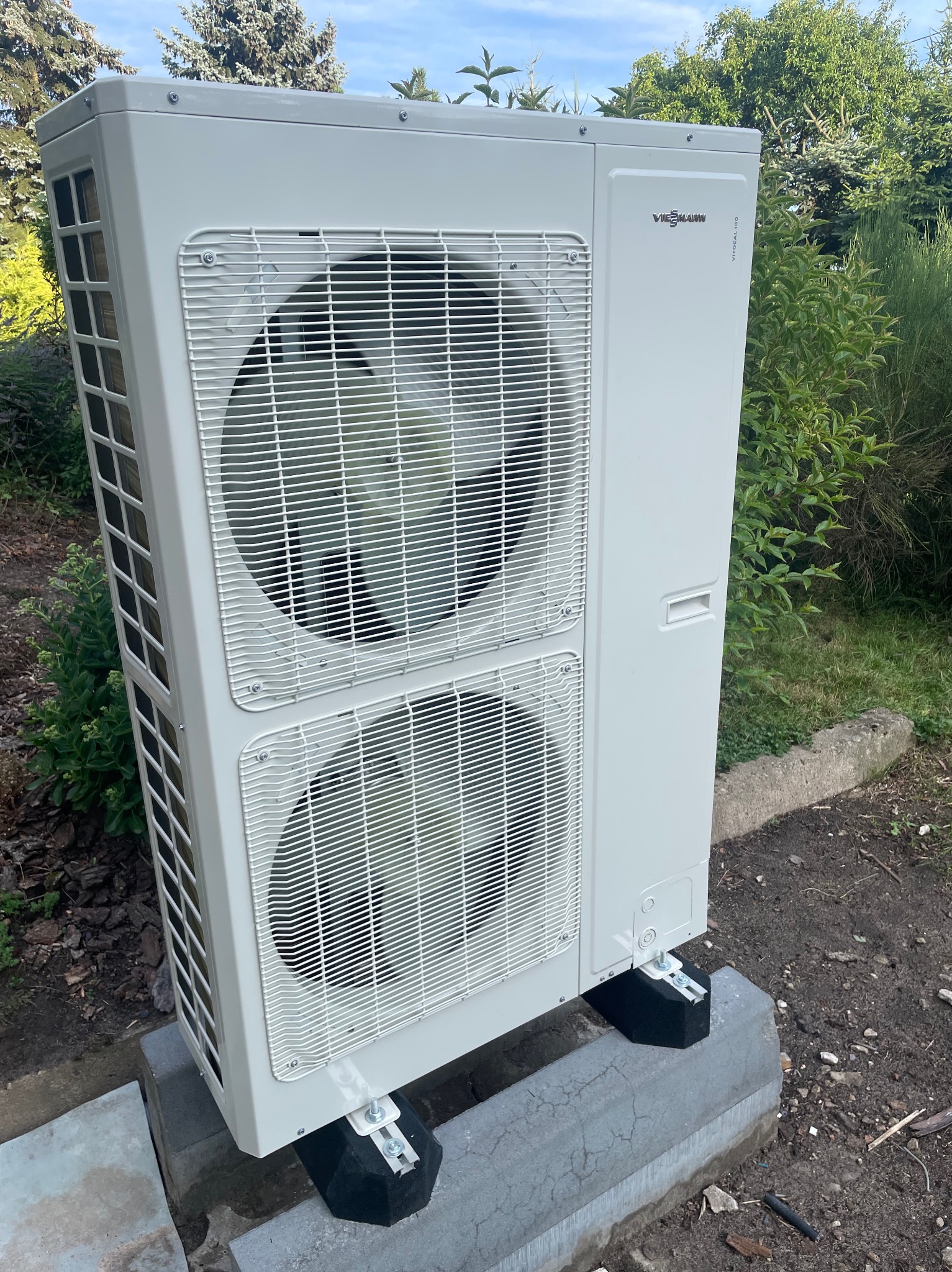 Biała pompa ciepła Viessmann Vitocal 100 umieszczona na betonowych blokach, otoczona zielenią w ogrodzie.