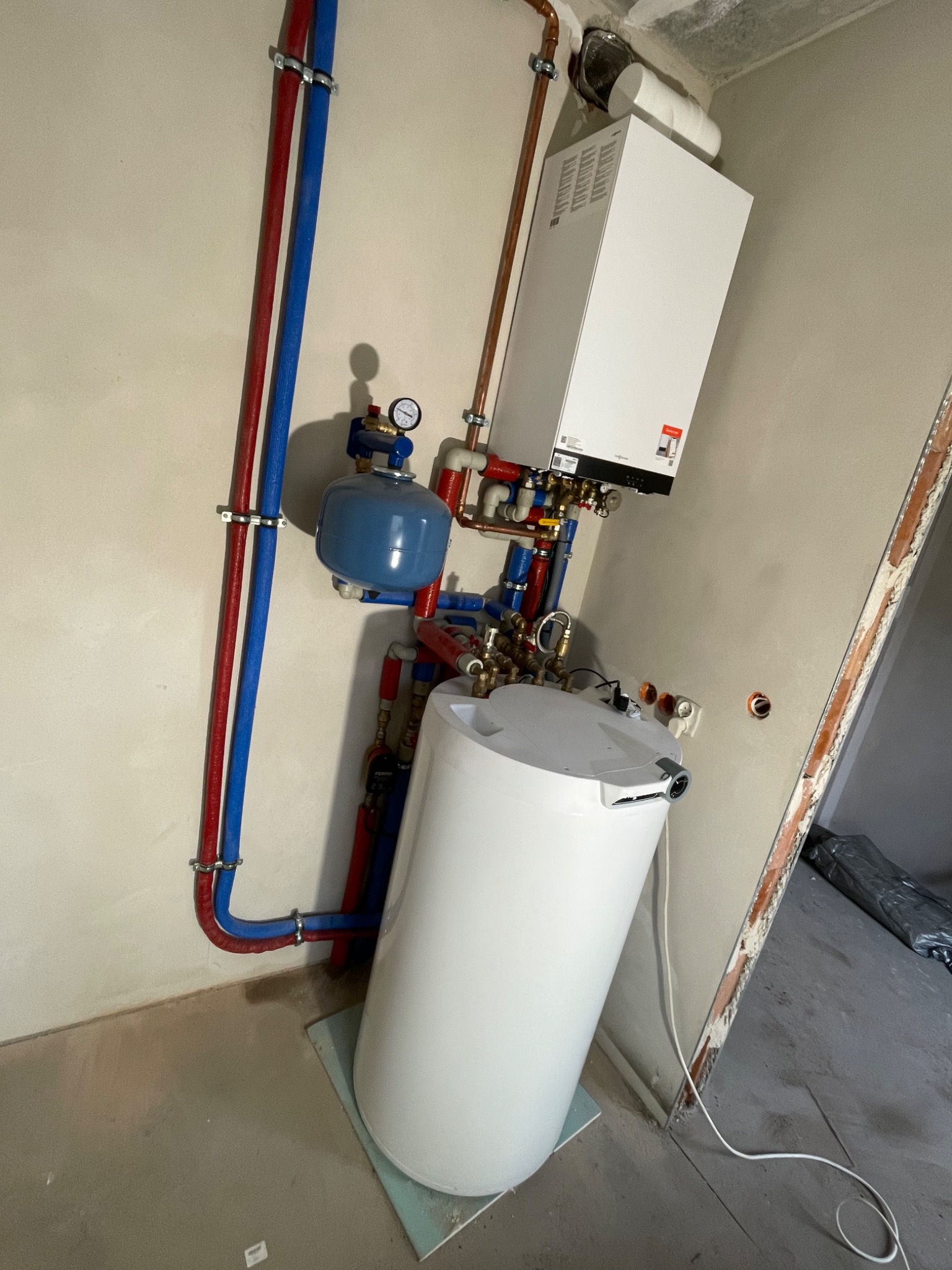 Zamontowany piec grzewczy Viessmann z zasobnikiem ciepłej wody, instalacja z rur miedzianych i PEX w kolorze czerwonym i niebieskim, widoczne manometry, surowy stan wykończenia pomieszczenia.