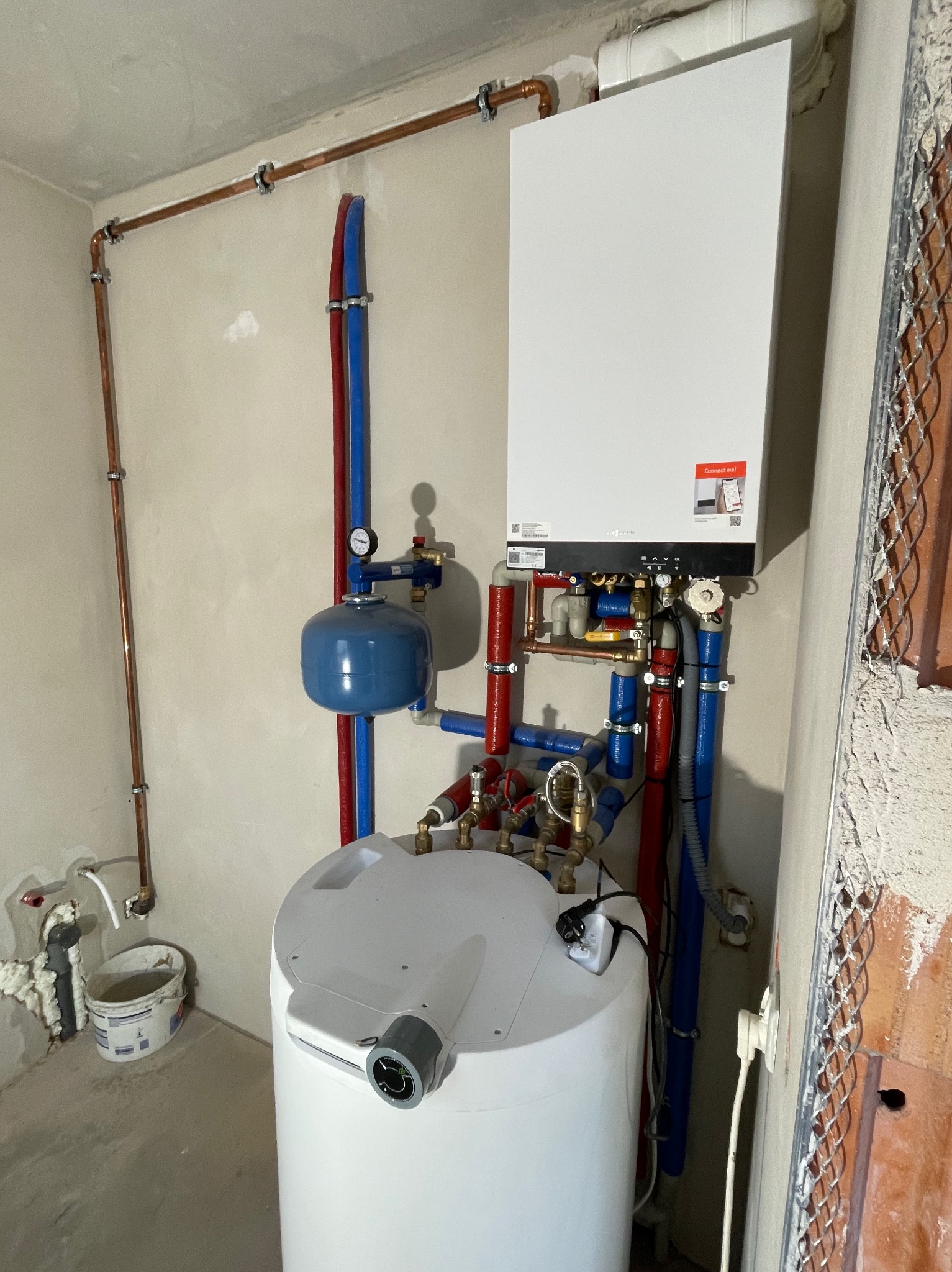Nowoczesny, biały piec grzewczy zamontowany na ścianie z widocznymi miedzianymi rurami, niebieskimi i czerwonymi przewodami, zbiornikiem wyrównawczym i zasobnikiem ciepłej wody w surowym...