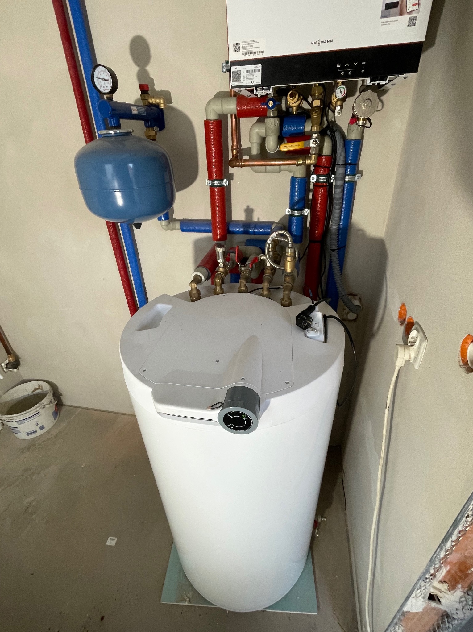 Biały zasobnik ciepłej wody użytkowej z podłączeniami rur miedzianych i plastikowych w kolorach czerwonym i niebieskim, zamontowany obok wiszącego kotła gazowego Viessmann i zbiornika wyrównawczego...
