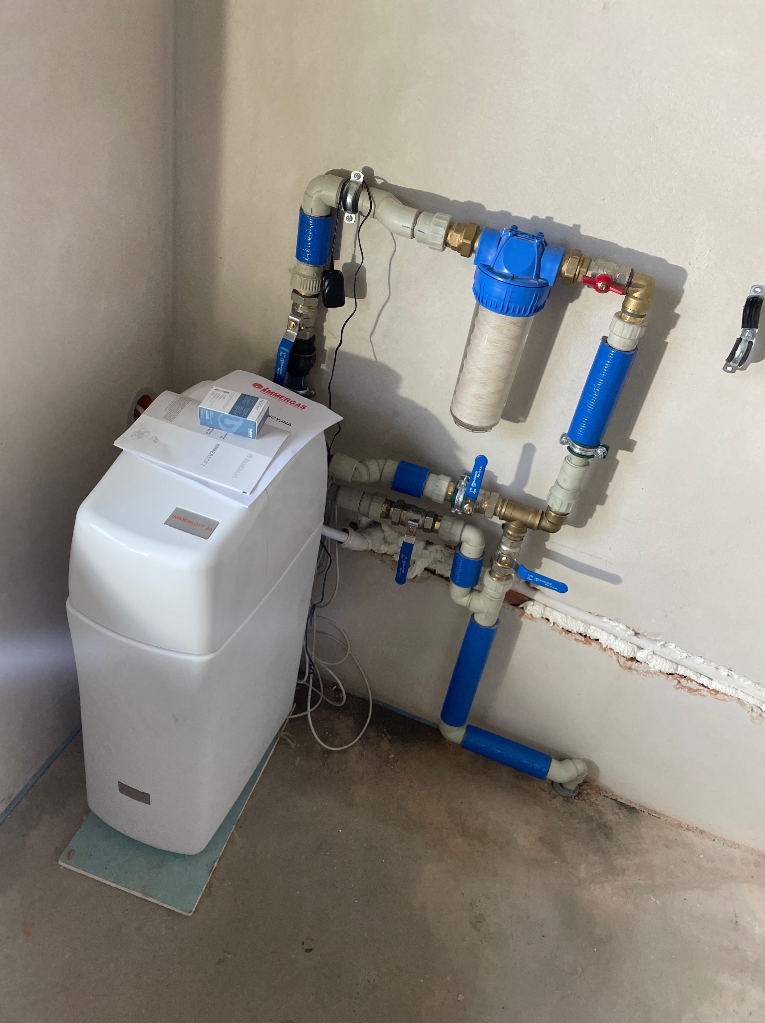 Biały zmiękczacz wody Immersoft 20 z niebieskimi rurami i filtrem mechanicznym w rogu pomieszczenia z surowymi ścianami. Na zmiękczaczu leżą dokumenty i pudełko.