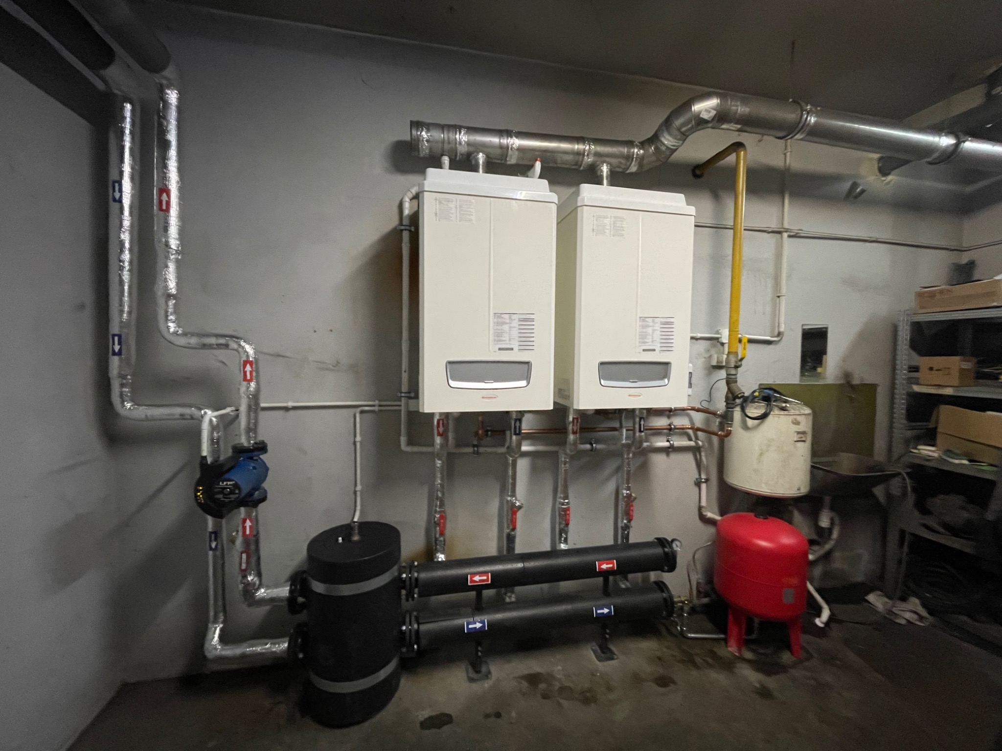 Dwa białe kotły grzewcze wiszące na szarej ścianie, połączone rurami z czarnym zbiornikiem wyrównawczym i czerwoną przeponą, instalacja w piwnicy.