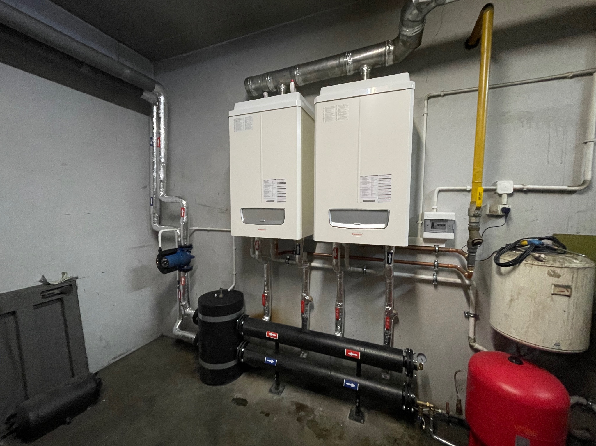 Dwa białe kotły grzewcze zamontowane na ścianie z widocznymi rurami miedzianymi, zbiornikiem wyrównawczym w kolorze czerwonym i instalacją wodną w piwnicy.