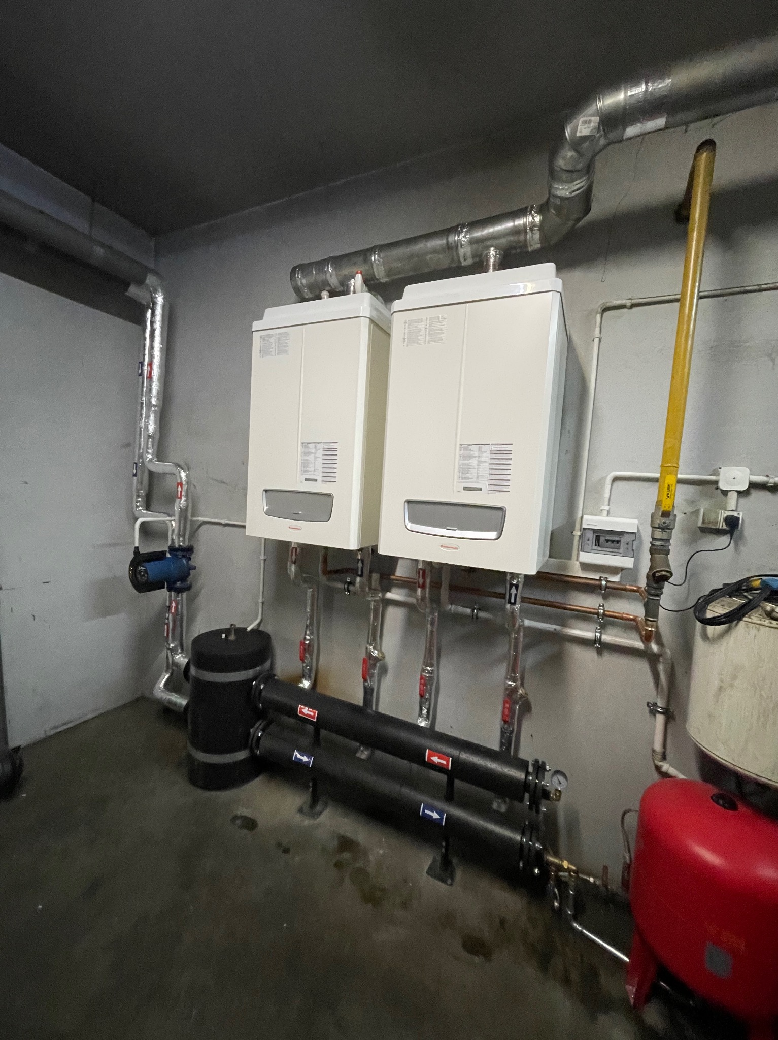Dwa białe piece grzewcze wiszące na szarej ścianie, z widocznymi rurami miedzianymi, czarnymi filtrami i czerwonym zbiornikiem wyrównawczym, w pomieszczeniu technicznym.
