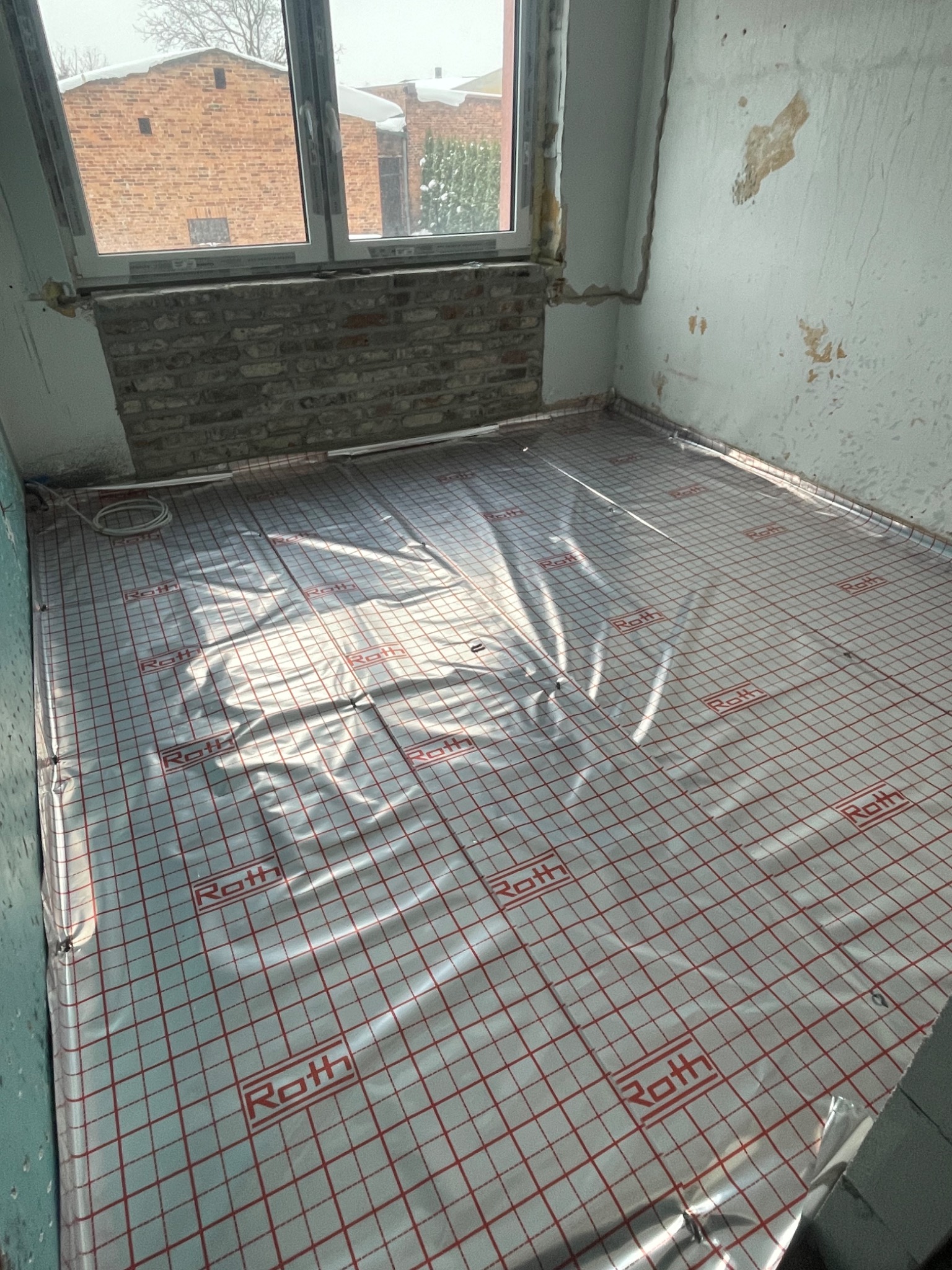 Przygotowanie do montażu ogrzewania podłogowego: folia systemowa Roth rozłożona na podłożu w surowym pomieszczeniu z oknem i fragmentem odsłoniętej cegły.