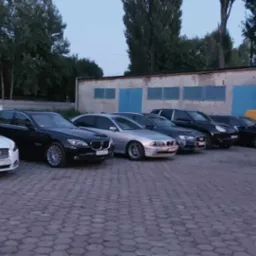 Rząd zaparkowanych samochodów różnych marek i modeli na tle budynku warsztatu z niebieskimi bramami, w tym biały Jaguar, czarny BMW i żółty van z otwartą maską.