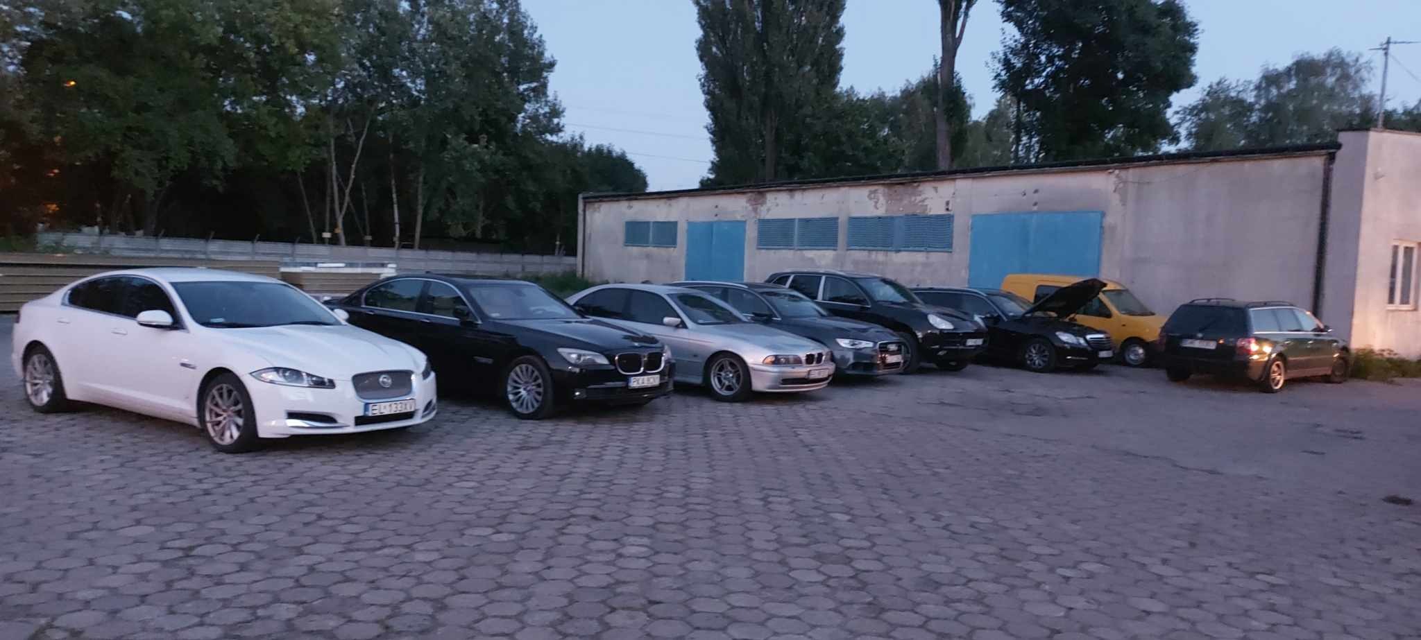 Rząd zaparkowanych samochodów różnych marek i modeli na tle budynku warsztatu z niebieskimi bramami, w tym biały Jaguar, czarny BMW i żółty van z otwartą maską.