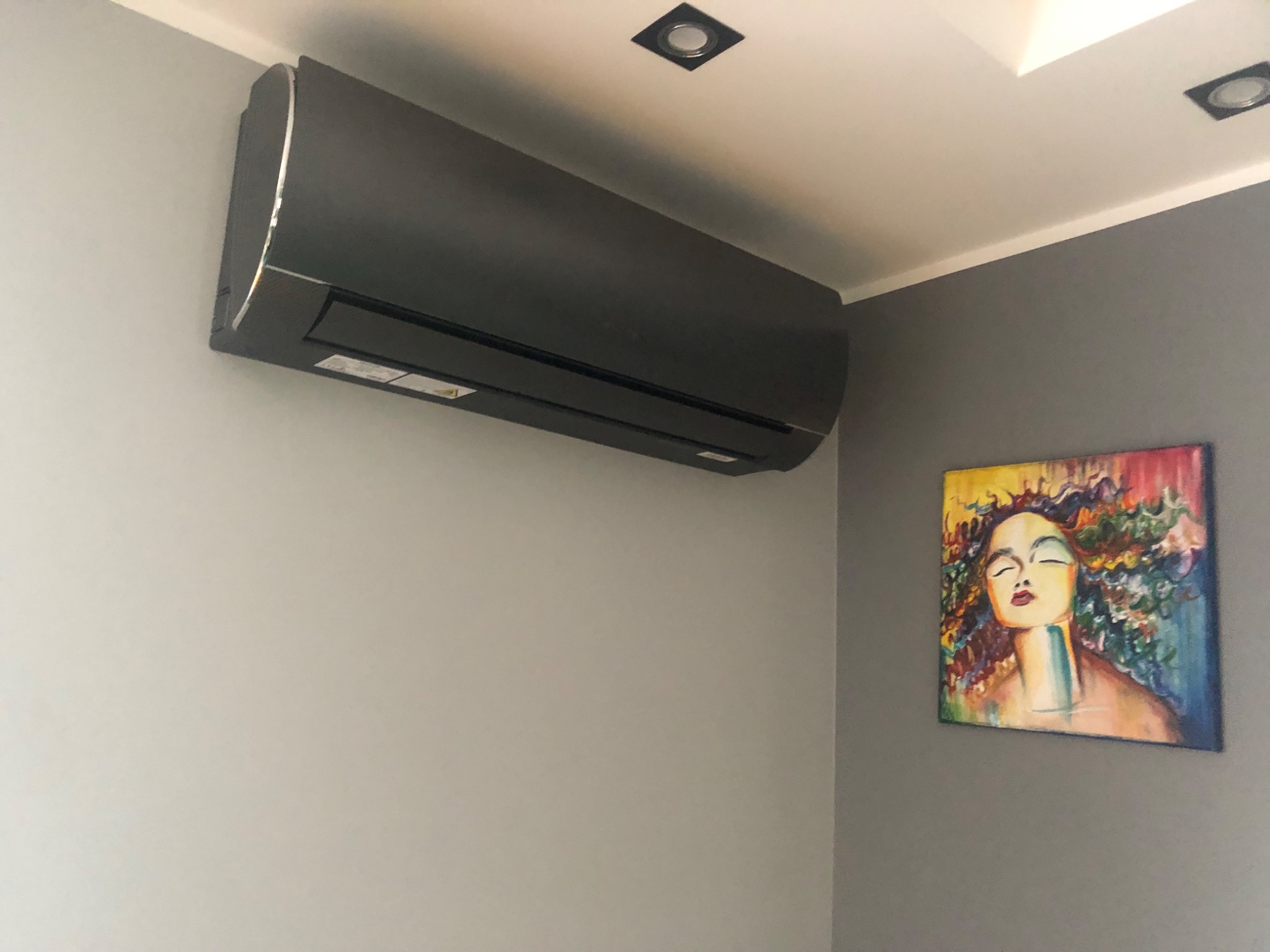 Ciemna jednostka klimatyzacji zamontowana na szarej ścianie w pomieszczeniu z obrazem kobiety. Widoczne oświetlenie sufitowe.