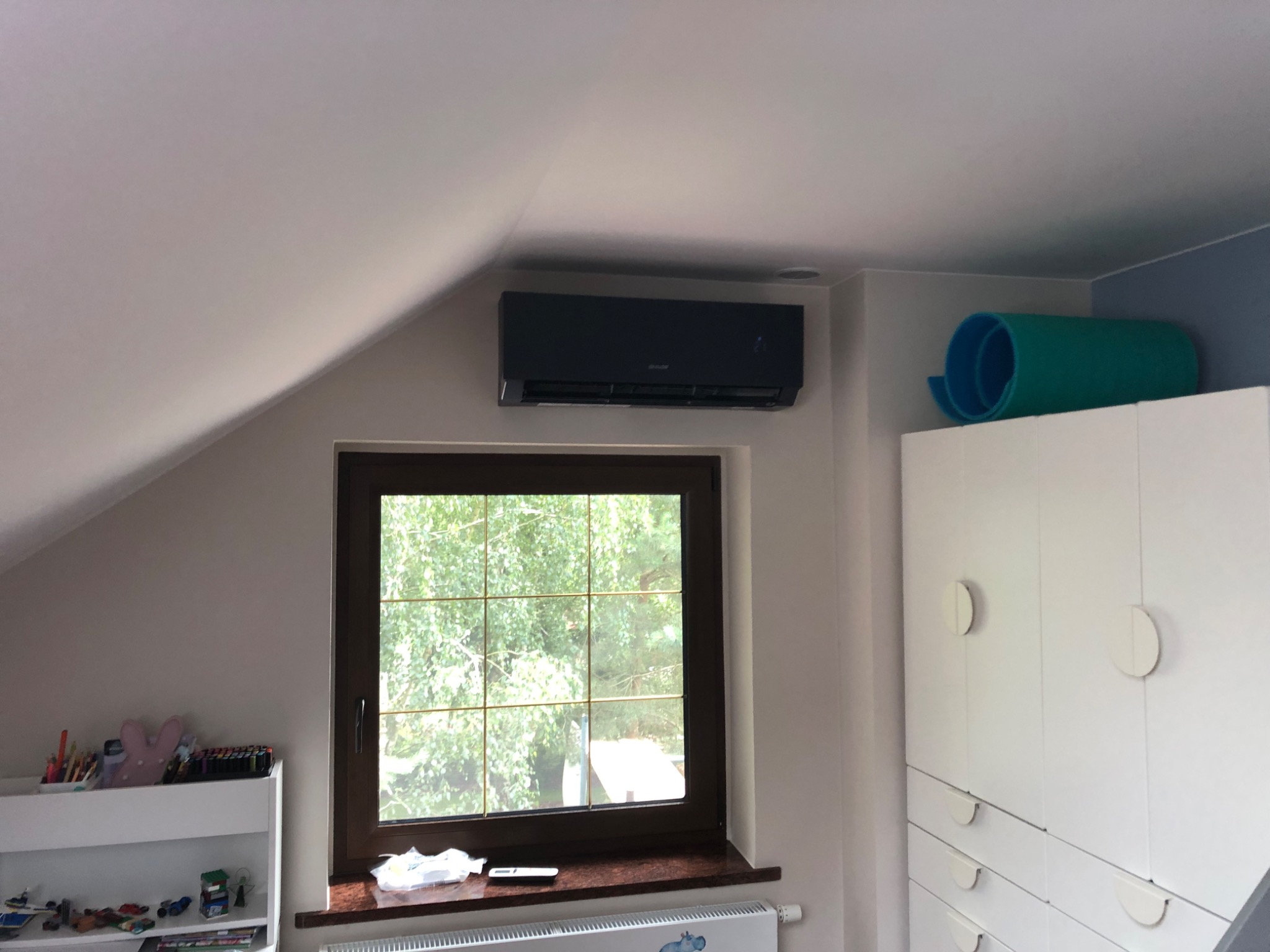 Czarny klimatyzator zamontowany nad oknem w pokoju na poddaszu, widoczny fragment białej szafy i grzejnika.