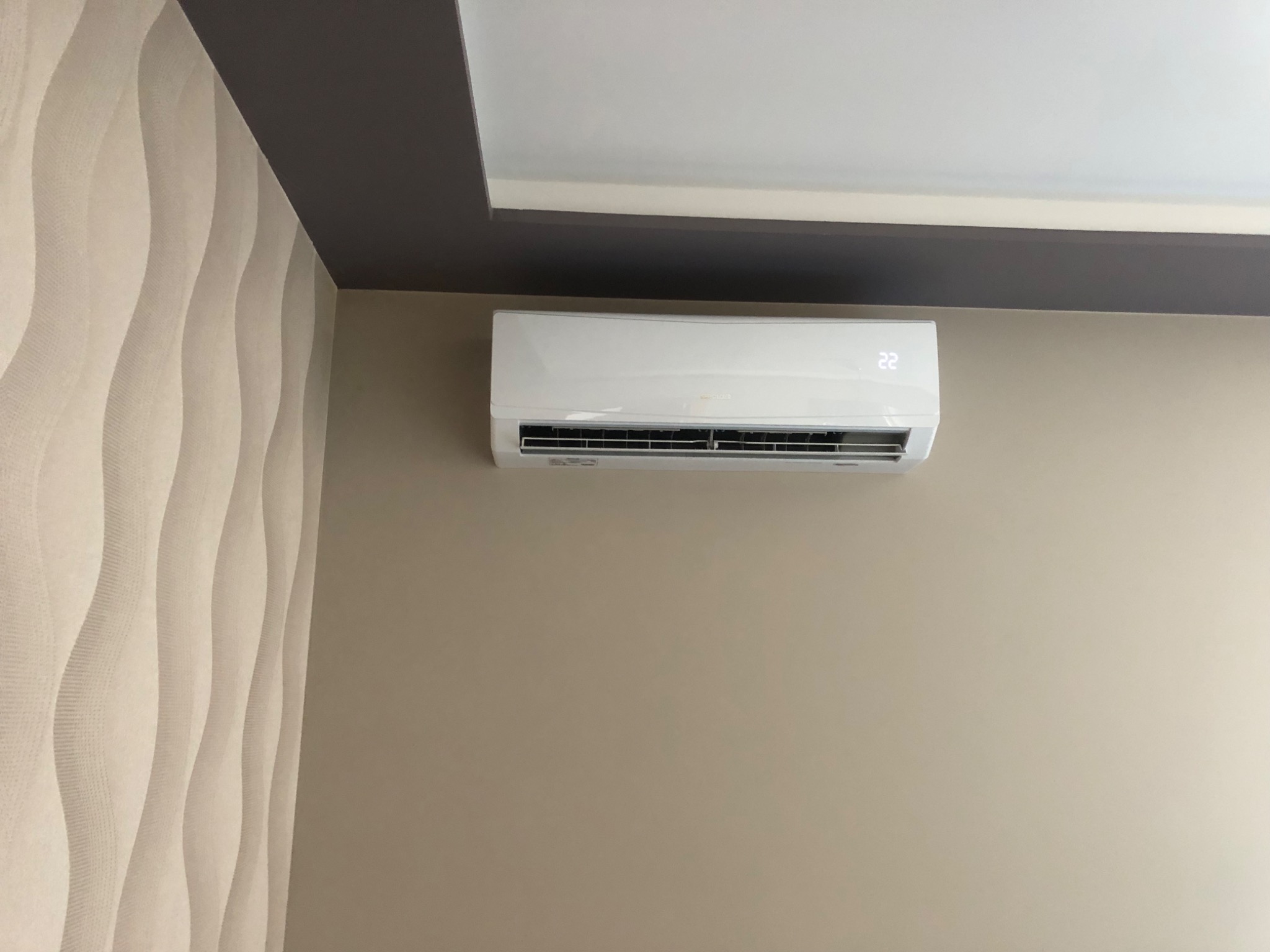 Biała klimatyzacja ścienna z wyświetlaczem wskazującym temperaturę 22 stopnie, zamontowana w jasnym pomieszczeniu z beżową ścianą i dekoracyjną tapetą w fale po lewej stronie.