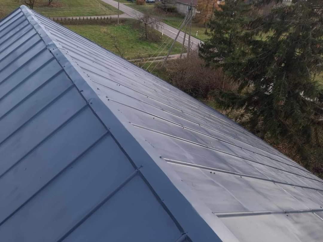 Nowo położony, szary dach z blachy trapezowej, widoczny z góry pod kątem, z elementami mocującymi; w tle drzewa i droga.