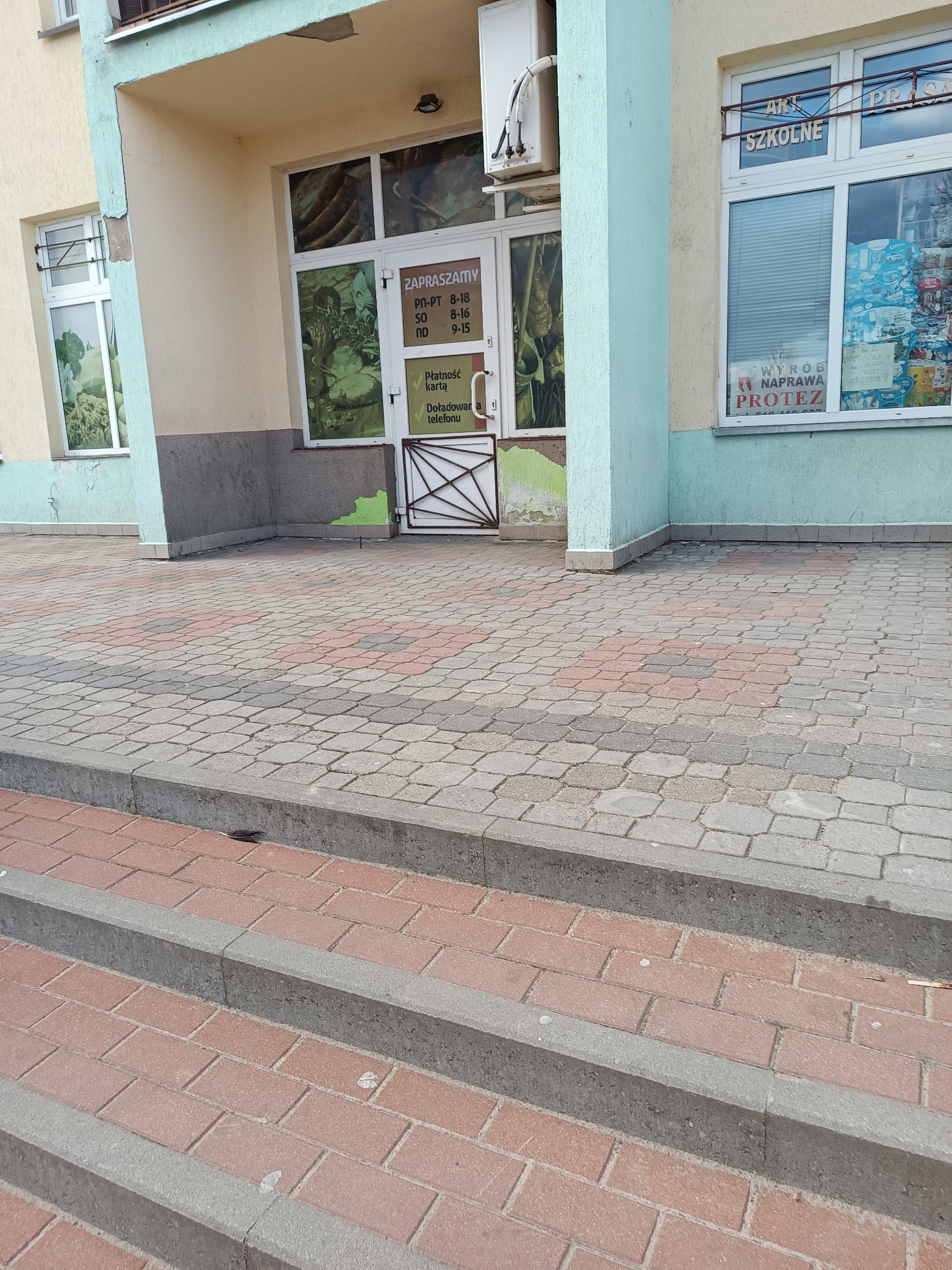 Fasada budynku usługowego z oknami i drzwiami wejściowymi, widoczne schody z kostki brukowej i tablica informacyjna na drzwiach.