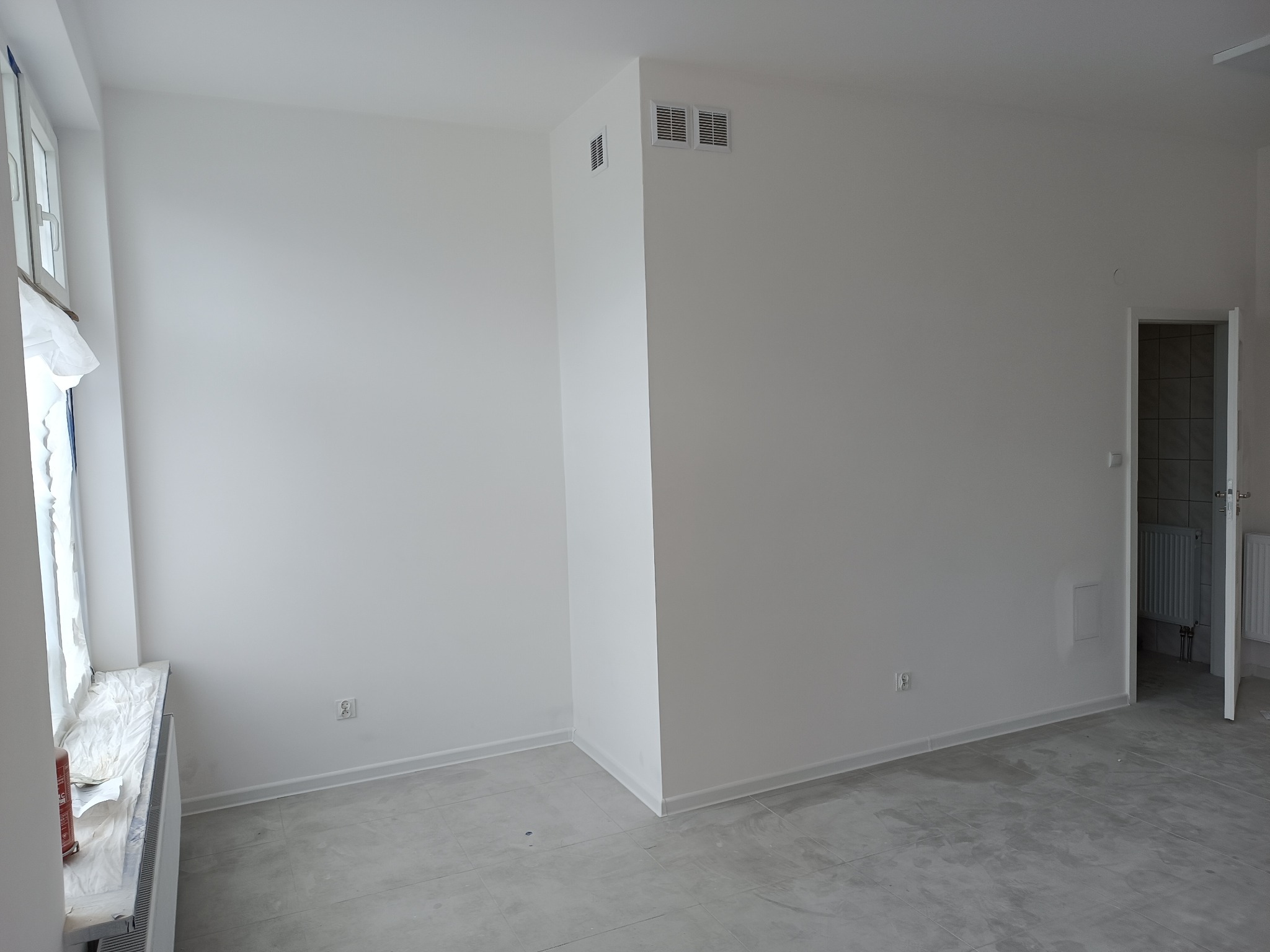 Puste, białe wnętrze po remoncie, z widocznym oknem zabezpieczonym folią malarską, kratkami wentylacyjnymi na ścianie oraz otwartymi drzwiami do łazienki z widocznym grzejnikiem.