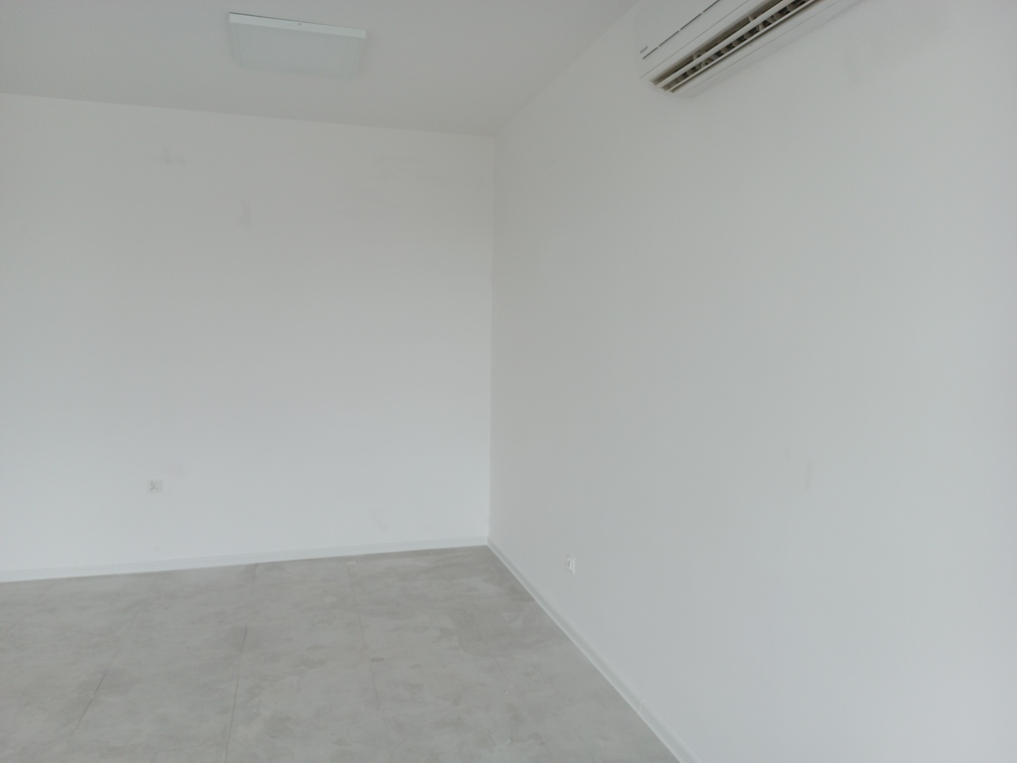 Pusty, nowo wyremontowany pokój z szarą podłogą z płytek, klimatyzacją na białej ścianie i prostokątną lampą sufitową.