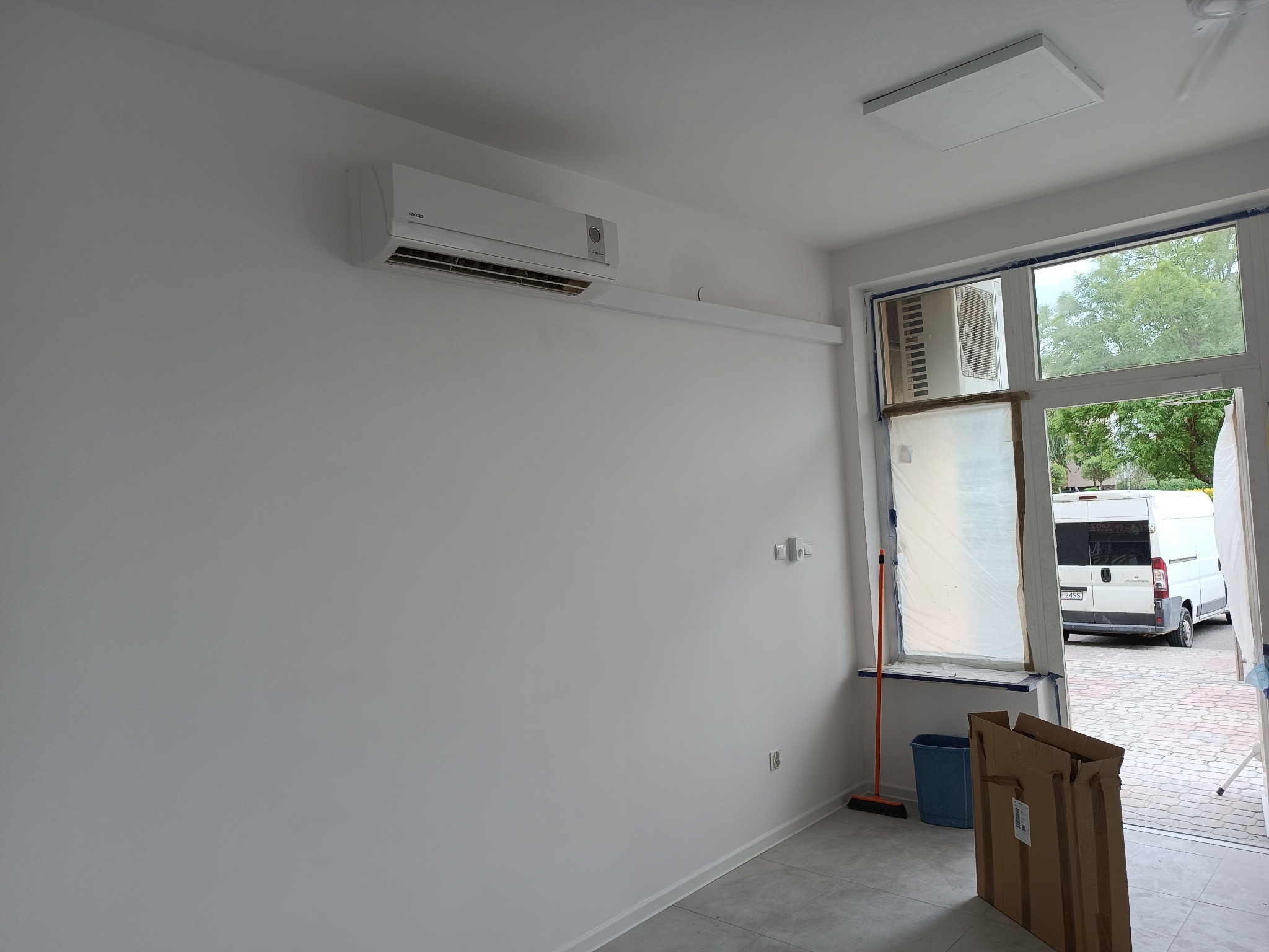 Wnętrze pomieszczenia po malowaniu, z zamontowanym klimatyzatorem na ścianie, okno zabezpieczone folią malarską, widoczny karton i wiadro na podłodze.