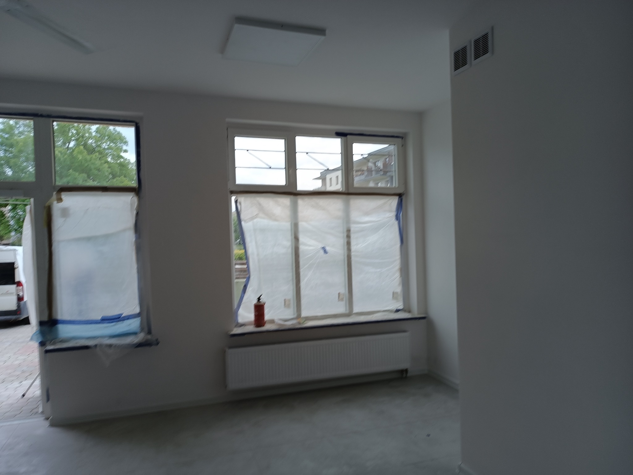 Wnętrze pokoju w trakcie malowania: okna zabezpieczone folią malarską, grzejnik pod oknem, gaśnica w rogu, ściany pomalowane na biało, widoczne kratki wentylacyjne.