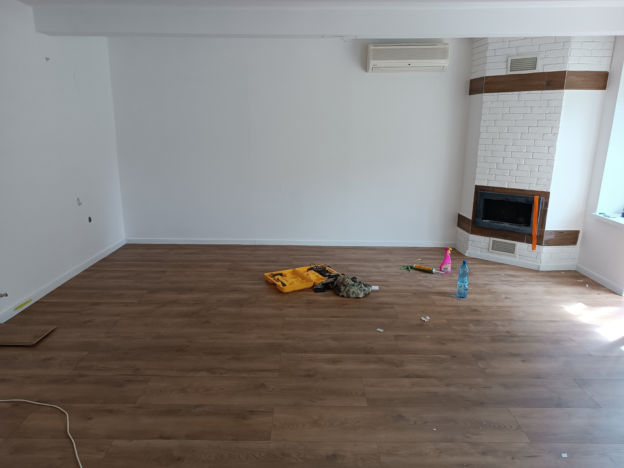 Pokój po częściowym wykończeniu: ułożona podłoga z paneli, białe ściany z widocznym klimatyzatorem i obudowanym kominkiem z białej cegły, narzędzia i akcesoria do montażu paneli podłogowych...