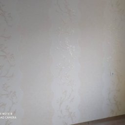 SW-DOM Wiktor Szymaniec - Jasna ściana z dekoracyjną tapetą w kwiatowe wzory, widoczne delikatne pionowe pasy z subtelnym tłoczeniem, ujęcie z dołu.