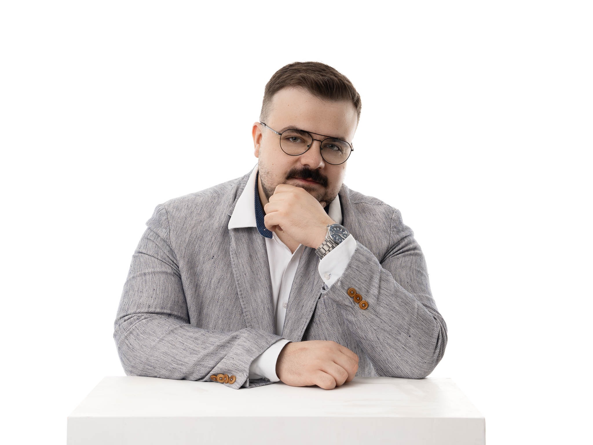 Mężczyzna w okularach, szarej marynarce w jodełkę i białej koszuli, siedzący przy białym stole, z ręką pod brodą i zegarkiem na nadgarstku.