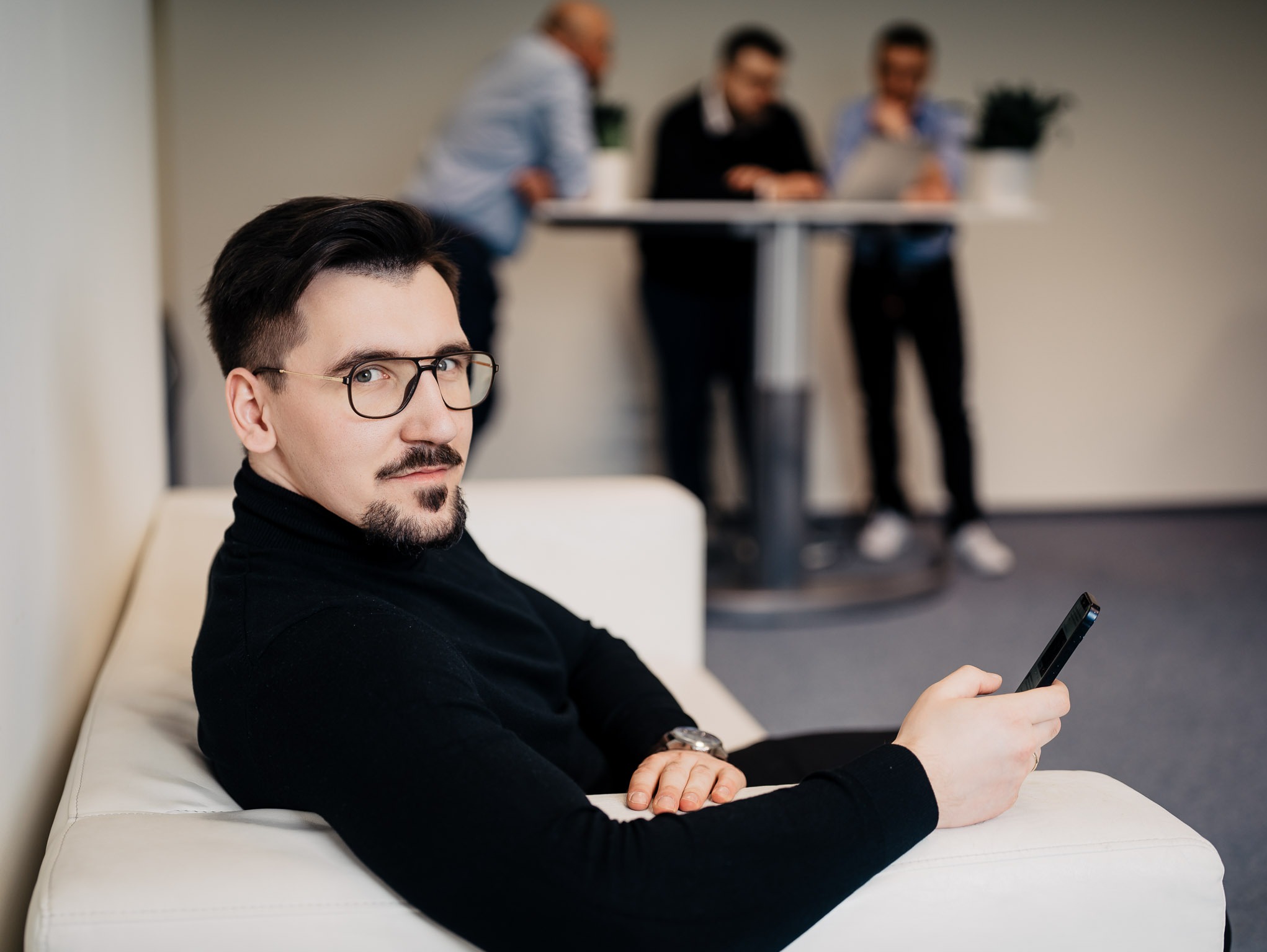 Mężczyzna w czarnym golfie i okularach siedzi na białej sofie, trzymając smartfon w dłoni; w tle widać grupę osób przy stole.
