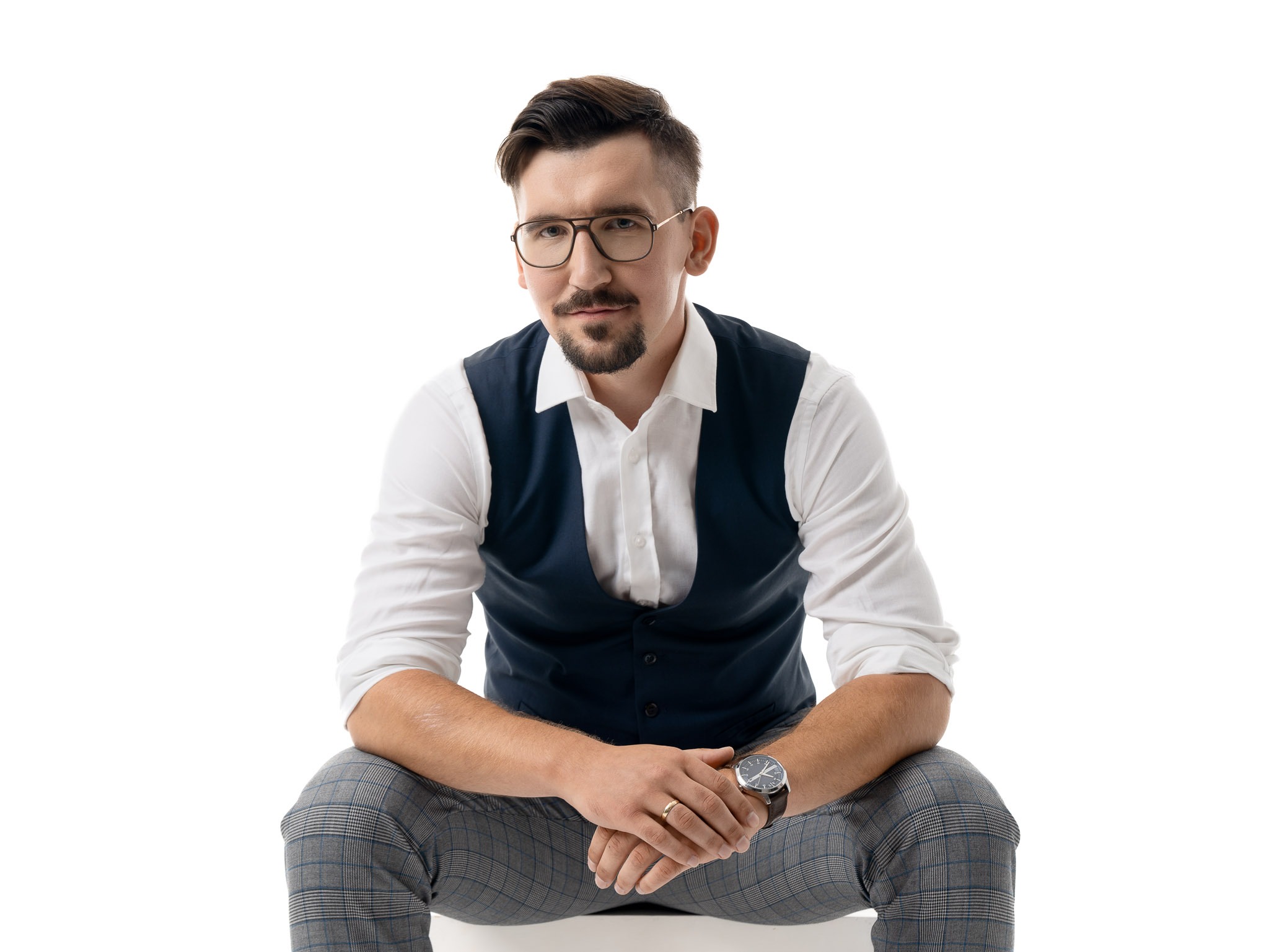 Mężczyzna z brodą i wąsami w okularach, białej koszuli, granatowej kamizelce i kraciastych spodniach, siedzący na białym tle, z zegarkiem na ręku i obrączką na palcu.