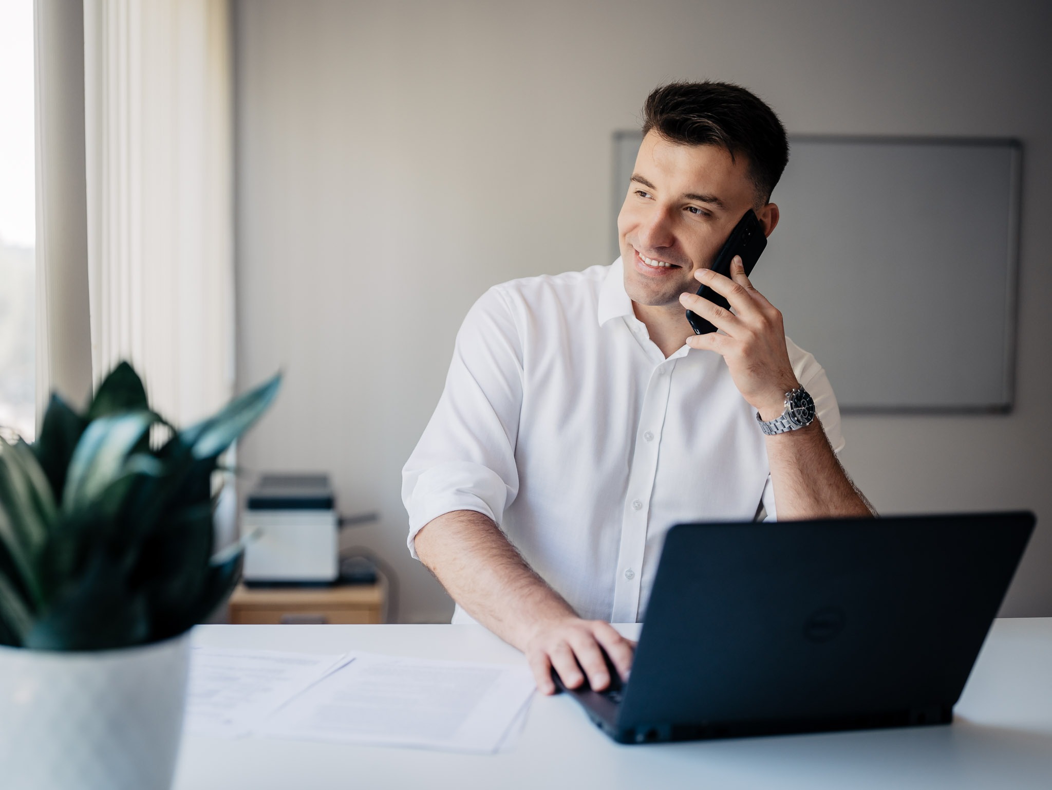 Uśmiechnięty mężczyzna w białej koszuli rozmawia przez telefon i pracuje na laptopie Dell, na biurku leżą dokumenty, w tle widoczne biuro z rośliną doniczkową.