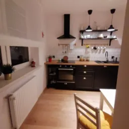 Wnętrze kawalerki z drewnianą podłogą, czarnymi szafkami kuchennymi, białymi płytkami na ścianie i białym krzesłem z żółtą poduszką.