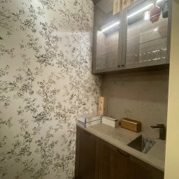 BDM Furniture - Niewielka kuchnia z ciemnymi szafkami, blatem z szarego kamienia i wzorzystą tapetą w kwiaty. Górne szafki z przeszklonymi drzwiczkami podświetlone LED, na górze drewniane skrzynki.