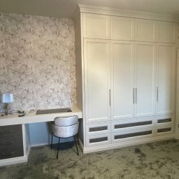 BDM Furniture - Biała szafa wnękowa z szufladami i biurkiem wbudowanym po lewej stronie, tapeta w tle z motywem roślinnym, srebrna lampka na biurku.
