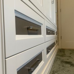 BDM Furniture - Biała szafa z szufladami i metalowymi uchwytami, widok z perspektywy podłogi na tle zielonego dywanu i białych drzwi.